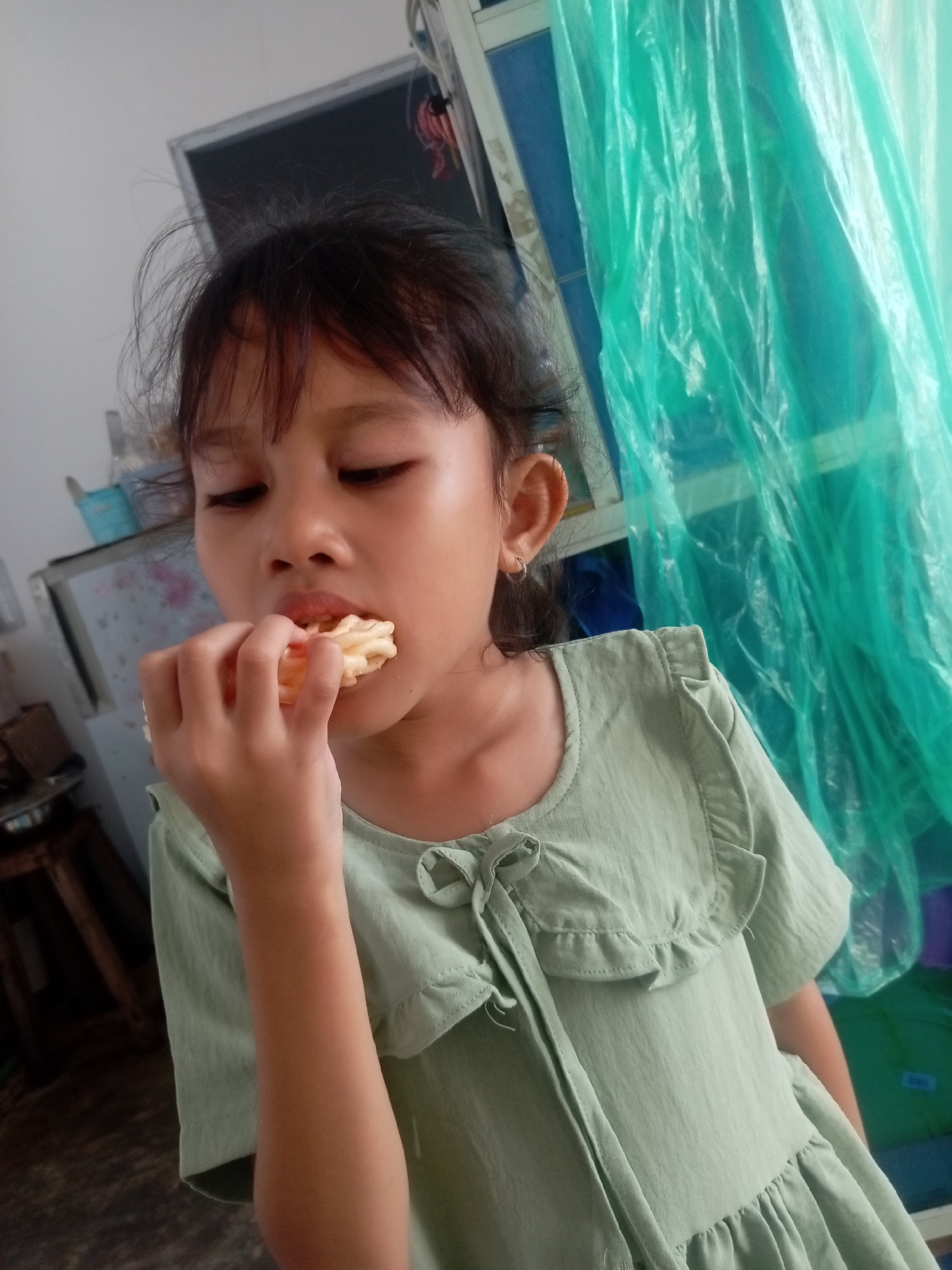 A young girl is eating a snack in Batam, Indonesia. She is wearing a light green dress with ruffled sleeves and a tie at the neck. The background shows a room with a refrigerator and a green plastic curtain. The lighting suggests it is daytime.