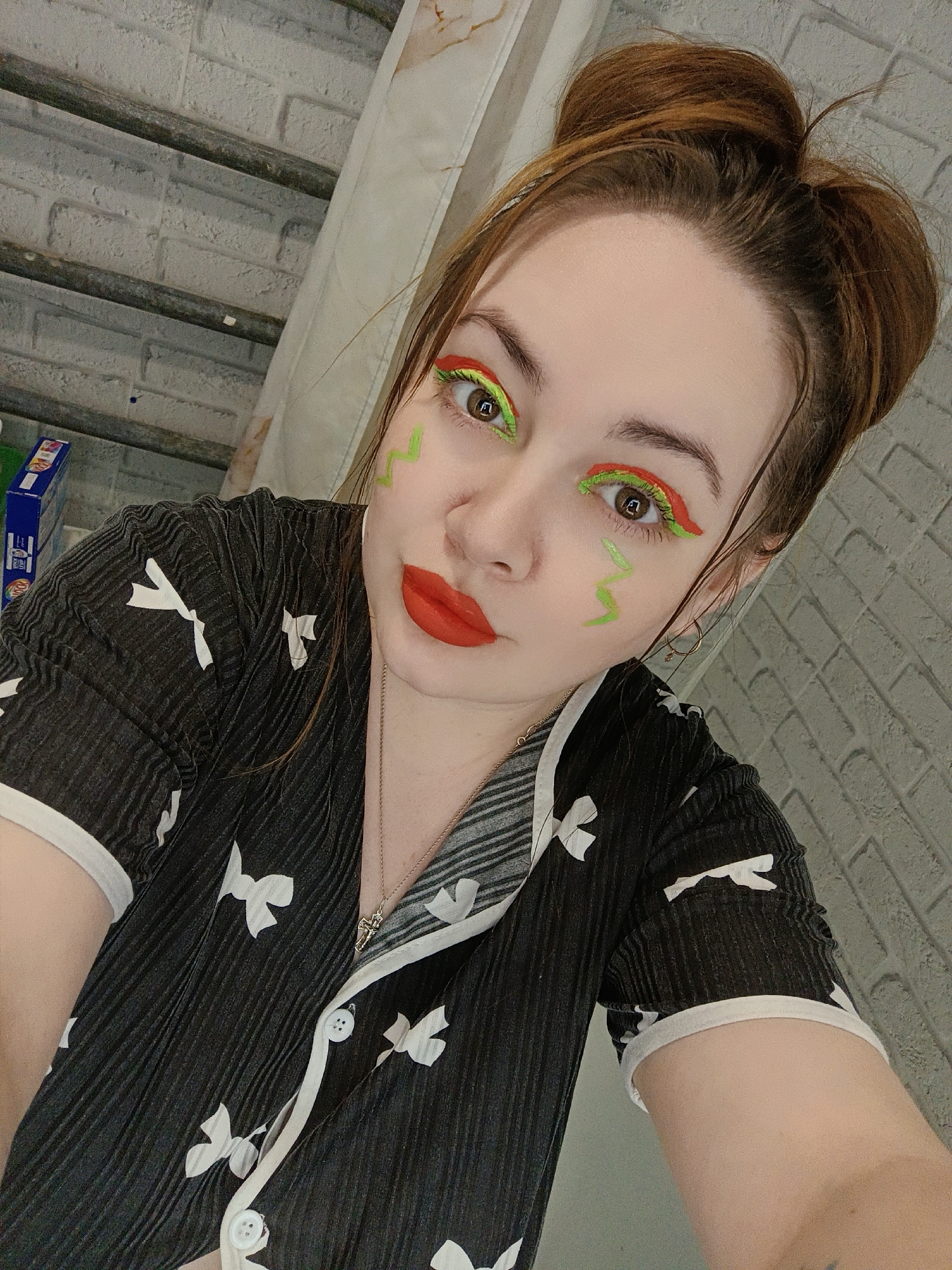 A close-up selfie captures a young individual indoors in Chaikovsky, Russia. The subject, likely female, has fair skin and brown hair pulled into a high, messy bun. Their face features striking and unconventional makeup: vibrant lime green and orange graphic eyeliner is applied to both upper and lower eyelids, and bright red lipstick adorns their lips. Two wavy lime green lines are drawn on their cheeks, extending from below the eyes. They look directly at the camera with a neutral, focused expression.
The individual is wearing a black short-sleeved top with a subtle ribbed texture, decorated with small white bow patterns and white trim along the collar and sleeves. A delicate gold chain necklace with a small charm is visible around their neck.
The background primarily features a white brick-patterned wall. In the upper left, dark horizontal elements, possibly beams or pipes, are visible against the wall. A light-colored, pleated fabric, resembling a curtain or drape, is partially seen on the right side of the frame. In the distant left background, a blue rectangular box with some faint, unreadable white text is present on a surface. The lighting suggests an indoor setting, likely well-lit.