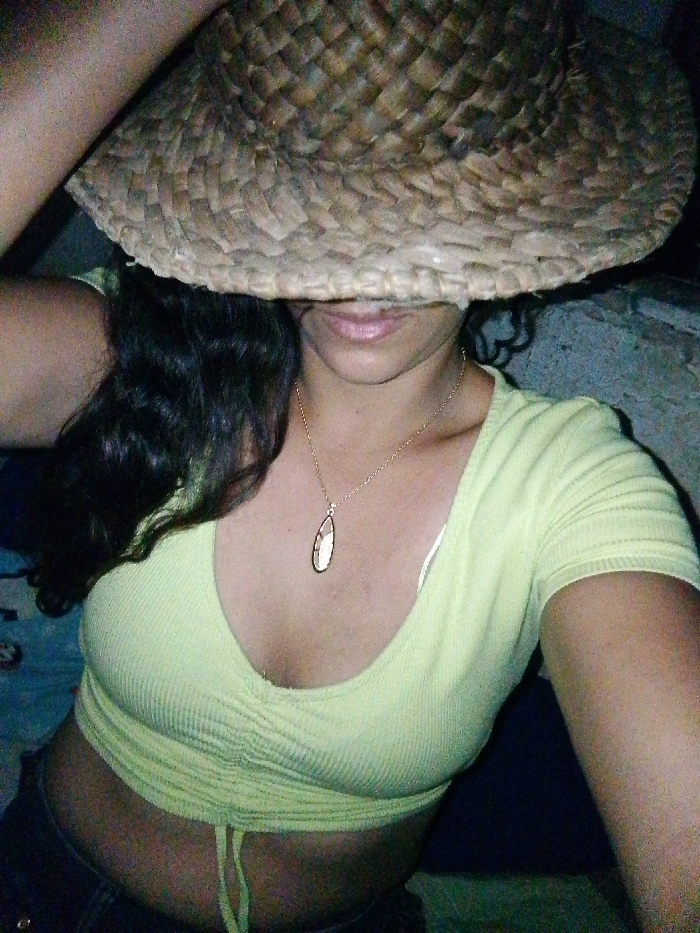 An image depicts an individual, identified as female, in Juan Griego, Venezuela. The subject's face is largely obscured by a woven straw hat with a wide brim, revealing only the lower lip and chin. Long, dark, wavy hair is visible on the subject's left side. The individual is wearing a light green, ribbed, V-neck crop top with short sleeves and a central drawstring detail, exposing the midriff. A delicate gold-colored chain necklace with an elongated, tear-drop shaped pendant, featuring a white or light-colored inlay, is visible around the neck. A white bra strap is visible on the right shoulder. Dark-colored trousers are partially visible at the bottom left. The subject's left arm is raised, with the hand positioned to hold the brim of the straw hat. The background consists of a dark, textured surface on the right, possibly stone or concrete, and a darker, undefined surface on the lower left, indicating a dimly lit environment.