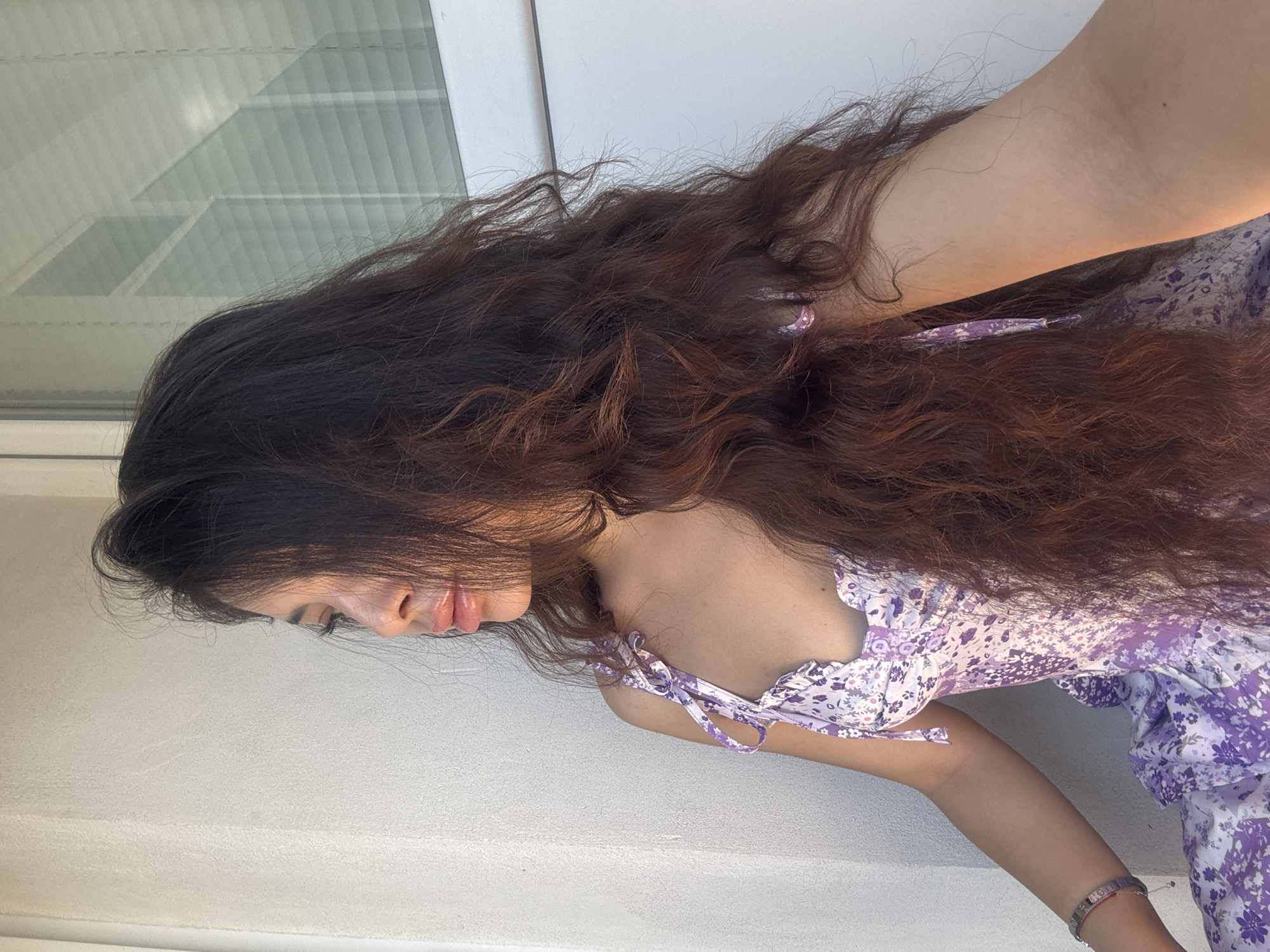A woman with long, dark, wavy hair poses in an outdoor setting, likely a balcony or terrace. She wears a light purple floral dress with ruffled details. Her face is partially visible, showing her lips and a glimpse of her eye. She is positioned against a background that includes a white wall and a window with blinds. The photo is taken from a slightly high angle. There is no evidence to suggest this photo was taken in Plovdiv, Bulgaria.