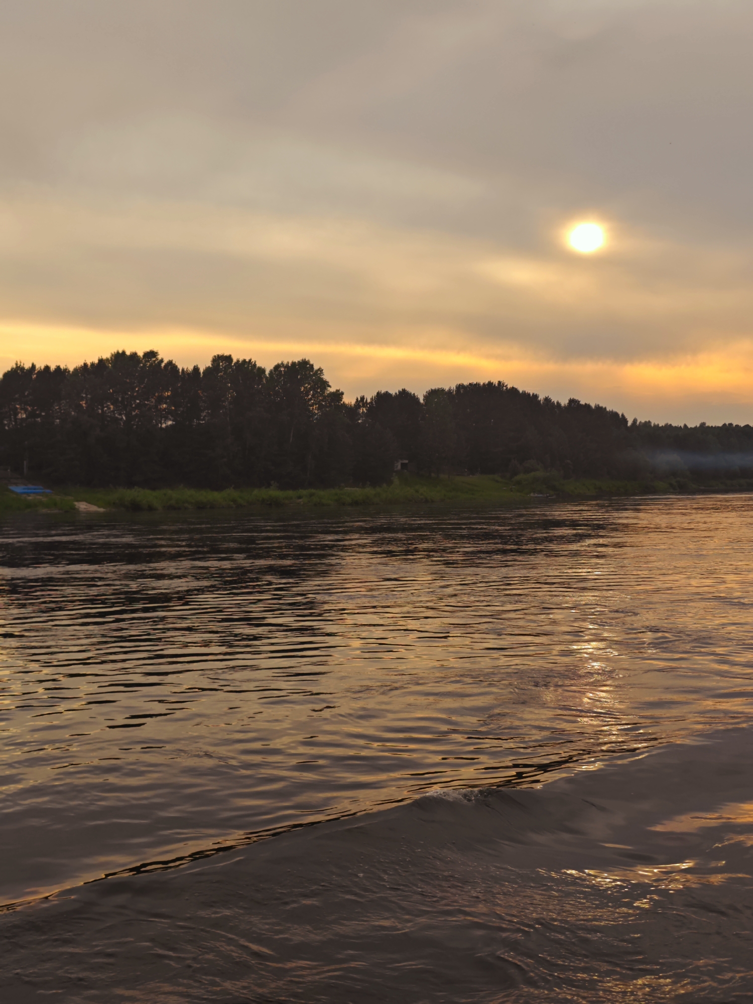 A photograph captures a sunset over a wide river, likely the Kama River, near the village of Yukseevo, Russia. The sky is a muted orange and yellow, with thin clouds partially obscuring the sun, which appears as a bright, hazy orb in the upper right quadrant of the image. The foreground is dominated by the river's surface, reflecting the warm hues of the sky and displaying ripples and a small wave generated by movement, possibly from a boat. On the far bank, a dense line of dark trees forms a silhouetted treeline against the horizon. Some green vegetation is visible at the base of the treeline. A faint plume of smoke rises from the treeline in the middle distance on the right side of the image. On the left bank, there are a couple of small, blue objects, possibly boats or tarpaulins, and a patch of light-colored sand. The overall impression is one of serene, natural beauty at dusk.