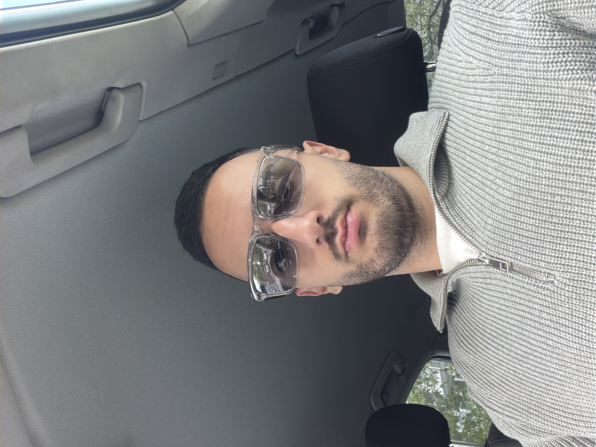 A man wearing clear-framed sunglasses and a grey zip-up sweater is seated in a car. The interior of the car is visible, including the ceiling, grab handle, and seat. The man has a short, dark haircut and a trimmed beard. The location cannot be identified as Plovdiv, Bulgaria, based on the visual information available.