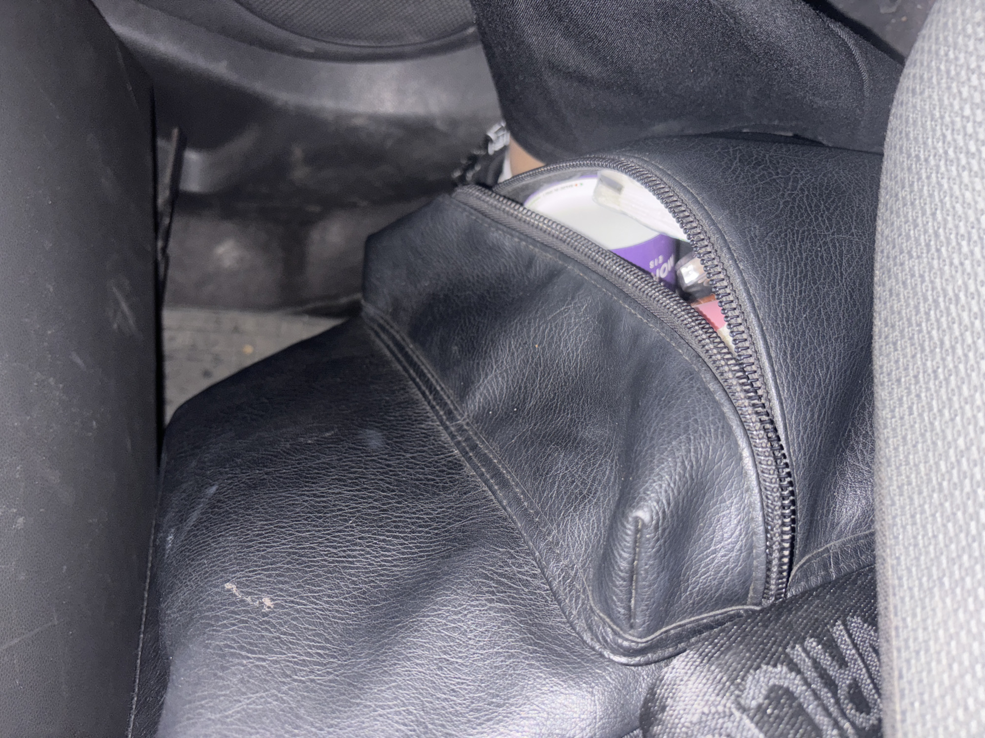 A black leather backpack is wedged between the seat and console of a car. The backpack's zipper is partially open, revealing a white object with purple accents inside. The seat is gray fabric. The car's interior is dark gray. No people are visible. The location, Kovačko Polje, Montenegro, is not discernible from the image.