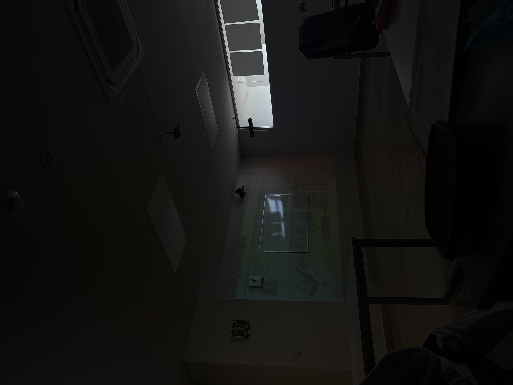 This media file captures a dim, underexposed interior view, likely a classroom or lecture hall, shot from a low angle looking upwards. Bright natural light streams through a large, rectangular skylight or window positioned on the upper right side of the frame, illuminating portions of the ceiling and upper walls.
On the ceiling, a white cassette-type air conditioning unit is visible, along with circular recessed fixtures, possibly lights or speakers. A significant portion of the wall in the mid-left displays a colorful presentation projected onto it. The projection features diagrams, illustrations, and text. A prominent vertical word in red reads "BIOLOGIA." Other partially decipherable text on the projection includes "COVID-19," "SINTOMAS" (Symptoms), and "PREVENCION," indicating the presentation's subject matter relates to biology, health, or disease. A reflective white board or frame is positioned over part of the projected image, causing a glare.
On a lower section of the wall to the bottom-left, a standard green emergency exit sign is mounted. It displays a white "running man" symbol and the Spanish text "SALIDA DE EMERGENCIA" (Emergency Exit) and "PUNTO DE REUNION" (Assembly Point).
In the bottom-right portion of the image, parts of dark, possibly upholstered, chairs and a dark metal table or desk frame are visible. A person's leg, clad in dark trousers, is also discernable at the very bottom right, suggesting someone is seated.
The room appears to be purposefully darkened to enhance the visibility of the projected image, despite the bright natural light from the skylight, indicating it is daytime. The Spanish text on the projection and the emergency sign aligns with the given location context of Puebla, Mexico.