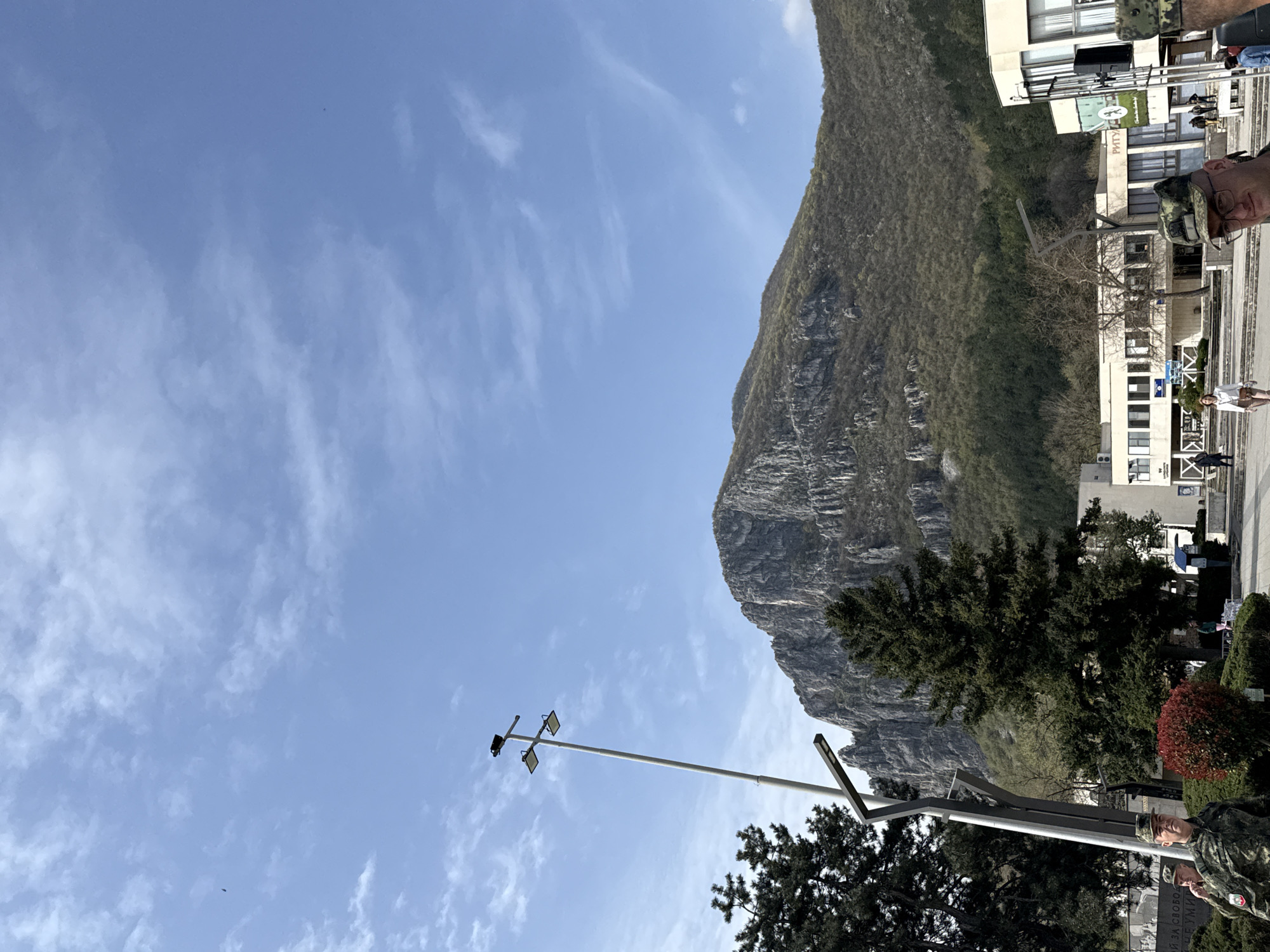 The image shows a daytime scene in Vratsa, Bulgaria. A large, rocky mountain, partially covered in green vegetation, dominates the background. A light blue sky with scattered white clouds is visible above the mountain. In the foreground are trees and a lamp post. Part of a building is visible to the right of the mountain. Two people in military-style clothing are standing near what appears to be a memorial or monument. The scene is oriented sideways.