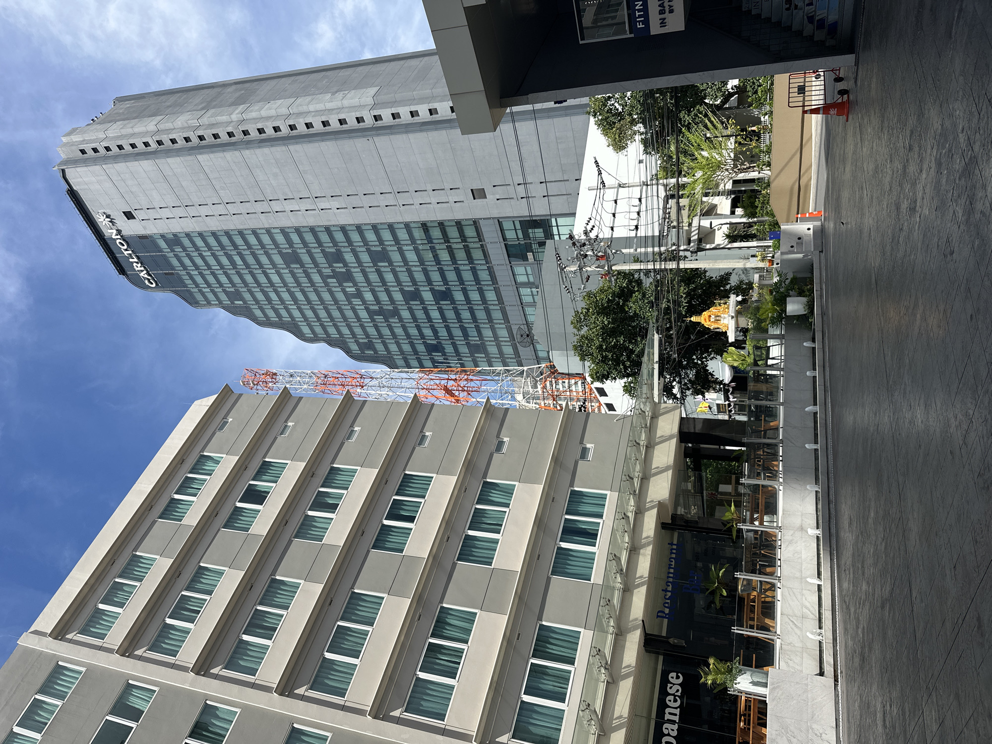 The image shows a street-level view in Bangkok, Thailand. Dominating the scene are two tall buildings. One is a grey, modern skyscraper with the name "CARLTON" visible on its side, reflecting the blue sky in its many windows. The other building has a light beige facade and a repetitive pattern of windows and protruding architectural features. A construction crane is visible above the buildings. The foreground features a section of sidewalk with a gray, patterned surface. A Japanese restaurant with outdoor seating is also in view. A small, golden Thai spirit house can be seen amidst some greenery in the background, adding to the local context. Overhead power lines and utility poles are present, a typical feature of Bangkok's urban landscape. An orange traffic cone sits on the sidewalk near a set of stairs. The weather appears to be sunny with a blue sky and some scattered clouds.