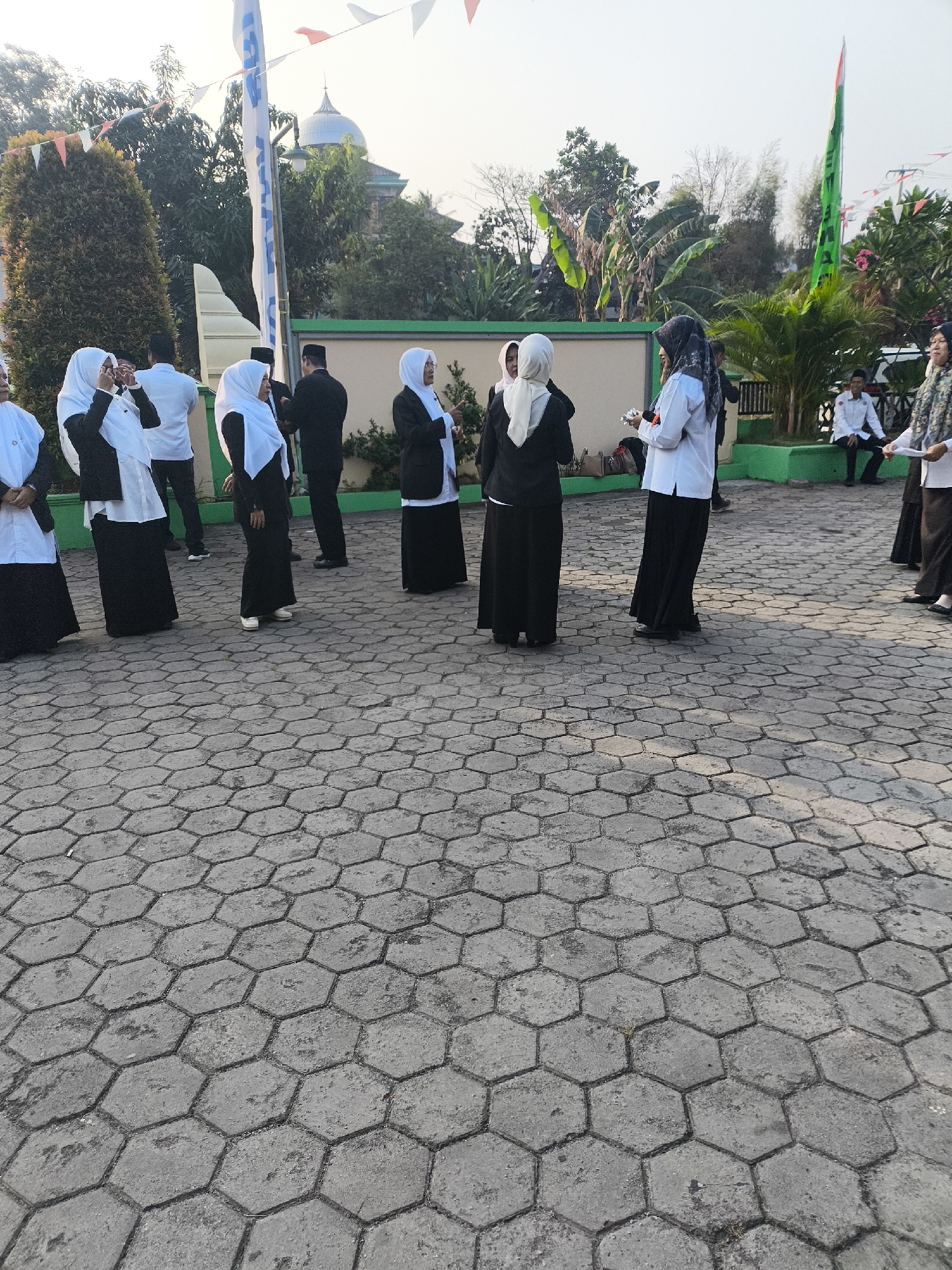 In Cilegon, Indonesia, a group of people, mostly women in black and white Islamic attire with hijabs, are gathered in an outdoor area with hexagonal paving stones. Some are conversing, while others appear to be listening or observing. In the background, a mosque with a dome is visible amidst lush greenery and trees. Flags, one predominantly green and white with Indonesian colors, are also present, suggesting a possible event or celebration. The overall atmosphere seems casual yet orderly, with the people positioned in various groupings on the paved ground.