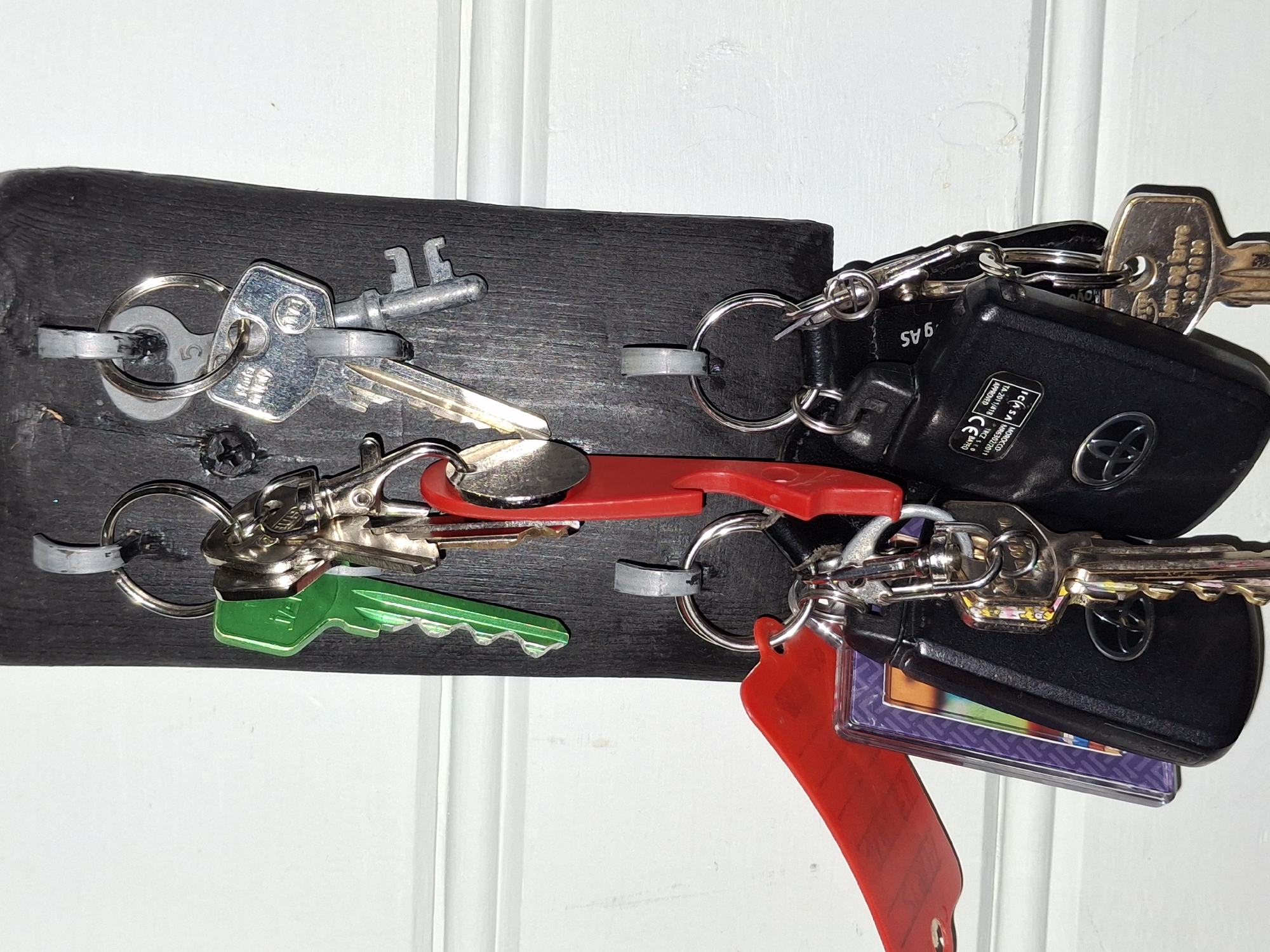 A key holder made of dark wood is mounted on a white wall in Åsgårdstrand, Norway. The key holder has multiple hooks, each holding one or more keys.
The keys are of various types and colors, including silver, green, and red. Some of the keys are attached to keychains, one of which has a red strap. Two of the keys are black car key fobs with a Toyota logo. One of the key fobs has a sticker with text including "MOROCCO" and "APPROVED." Another key has text that appears to be "jern og brøve" followed by numbers.
The scene is well-lit, and the details of the keys and key holder are clearly visible. The background is a white wall with some subtle texture.