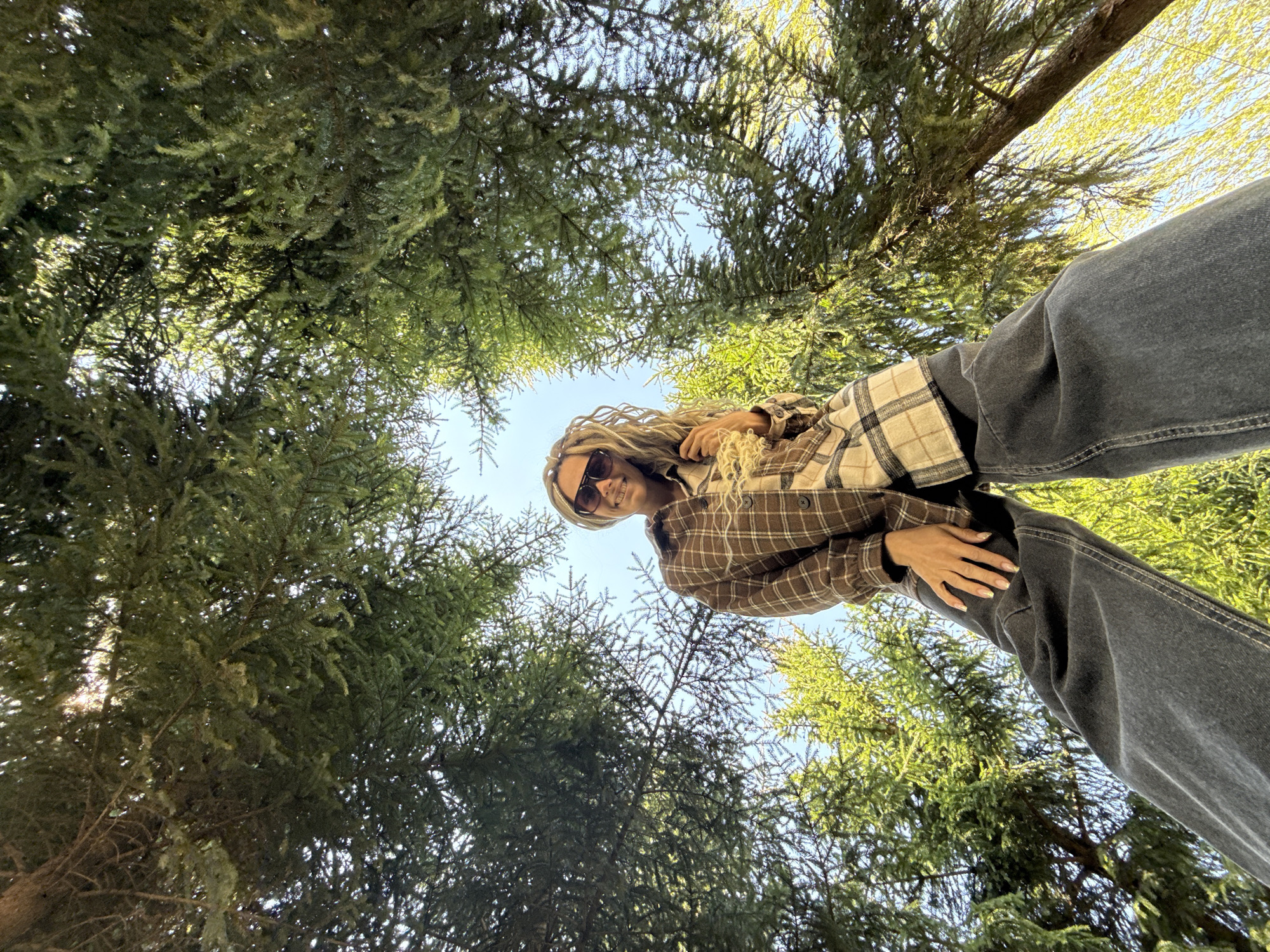 A low-angle shot shows a woman standing outside among trees. Her head, upper body, and part of her legs are visible in the image. She has blonde, dreadlocked hair and wears sunglasses, a brown plaid shirt, and dark jeans. Her right hand rests on her right thigh. Above her, a canopy of green trees fills much of the frame, allowing glimpses of the blue sky. The location is presumed to be a natural area within the city of Unknown, Unknown.
