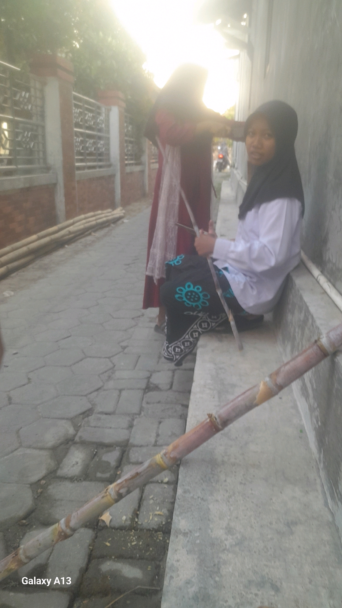 In Ngelo, Indonesia, two young women are featured in a street scene bathed in bright sunlight. The woman on the right, wearing a black hijab and a white shirt, is seated on a concrete ledge, looking towards the camera. She is holding a long, thin stick, possibly sugarcane, across her lap. Her dark skirt is adorned with a blue floral pattern and white geometric designs. To her left, another woman, partially obscured, wears a maroon hijab and a long, patterned maroon and white garment. She is standing and appears to be interacting with the seated woman, possibly handing her the stick. In the foreground, a piece of sugarcane lies diagonally across the paved walkway, which is made of hexagonal stone tiles. Piles of bamboo poles are stacked against a brick wall with a decorative metal fence to the left of the frame. The environment suggests an outdoor setting in a residential or commercial area, with a building wall forming the backdrop on the right.