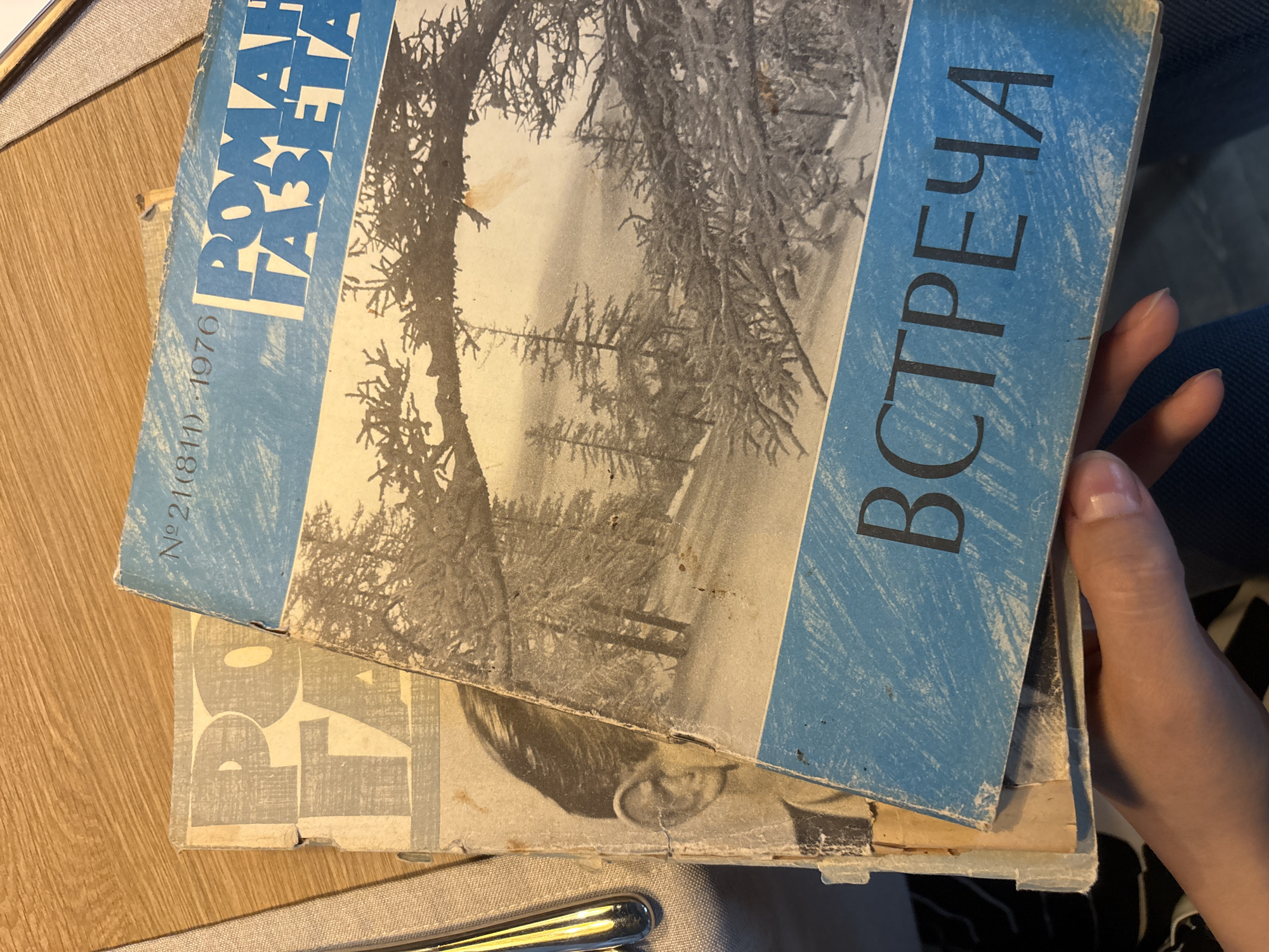 The image shows two vintage magazines stacked on a wooden table. The top magazine is blue with a black and white winter forest scene on its cover. The text "РОМАН ГАЗЕТА" is prominently displayed in large blue block letters. Above this, smaller text reads "№ 21(811) 1976". The bottom magazine, partially visible, has a pale cover with large grey block letters that appear to spell "PO FA". A portion of a person's ear and temple, belonging to a portrait on the bottom magazine, is visible. A person's hand is holding the bottom edge of the magazines. The scene is illuminated by natural light. The location is identified as Novaya Usman', Russia.
