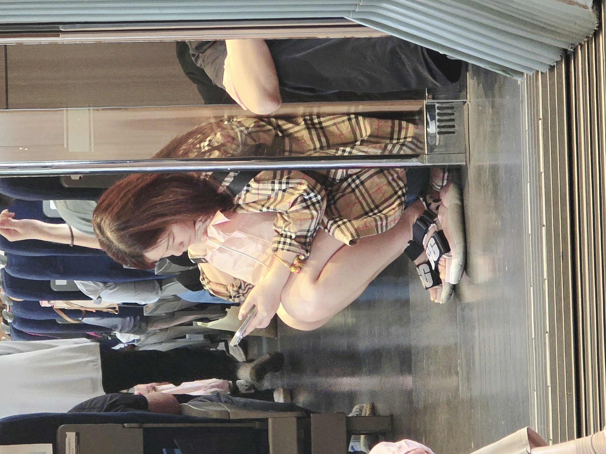 A young woman with reddish-brown hair is seen squatting inside a train car, looking down at her cell phone. She is wearing a plaid jacket over a pink top and blue shorts. Her feet are clad in chunky platform sandals with black straps. The train car has blue seats visible in the background, and the floor appears to be made of polished concrete. To her right, a person in a black shirt and pants is visible. The scene is captured from a low angle, looking through a glass partition.