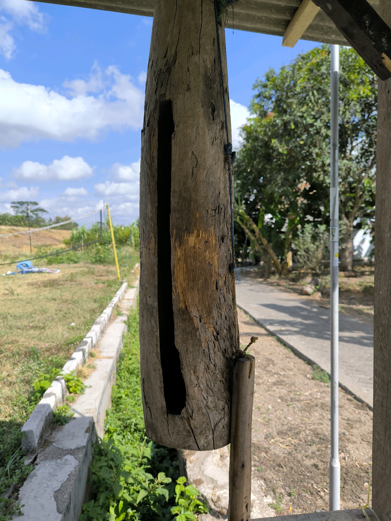 The image captures a close-up of a hollowed-out wooden object, suspended by a rope, in Karanganyar, Indonesia. The wood is weathered and appears old, with a long, dark cavity running vertically along its surface. A smaller piece of wood is attached to the bottom of the main object. The background reveals a sunlit outdoor scene with a blue sky dotted with white clouds. Green foliage, trees, a paved walkway, and a volleyball net are visible in the distance, suggesting an outdoor recreational area or a rural setting. The overall impression is of an object that might be a traditional instrument or a decorative piece in a natural environment.
