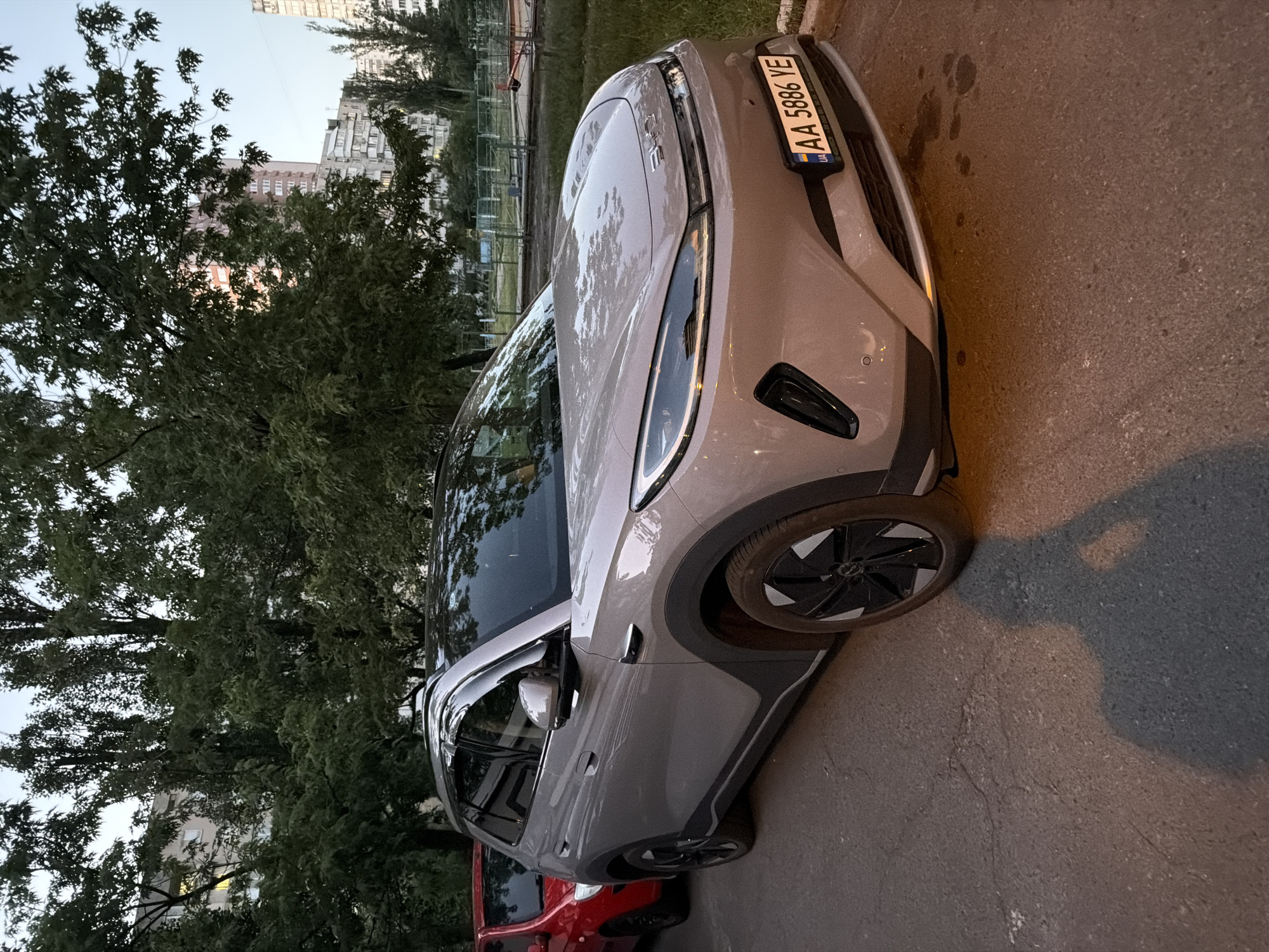 A light gray BYD Yuan Plus electric vehicle is parked on an asphalt surface next to a red car. The BYD has a black roof and a license plate that reads "AA 5886 YE". The car is angled towards the viewer, with its front right wheel visible. In the background, there are trees, apartment buildings, and a playground with a fence. The lighting suggests it is late afternoon or early evening. A shadow of a person holding a camera is visible on the asphalt in the foreground.