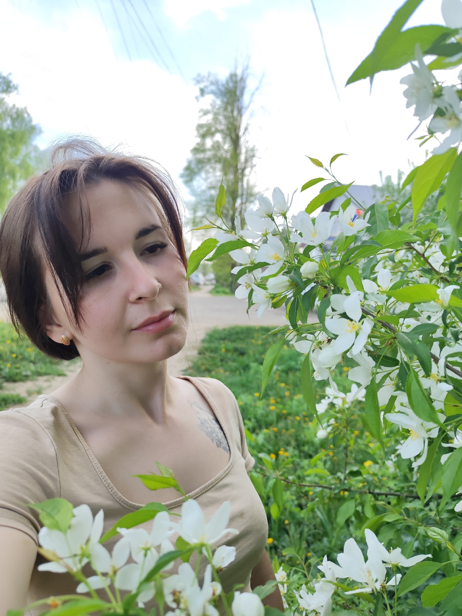 A woman stands outdoors next to a flowering tree. The woman has short brown hair and is wearing a tan-colored t-shirt. A tattoo is visible on her upper chest. The tree is covered in white flowers with yellow centers and green leaves. The background includes a street, some houses, trees, and a blue sky. The location is Фоки, Russia.