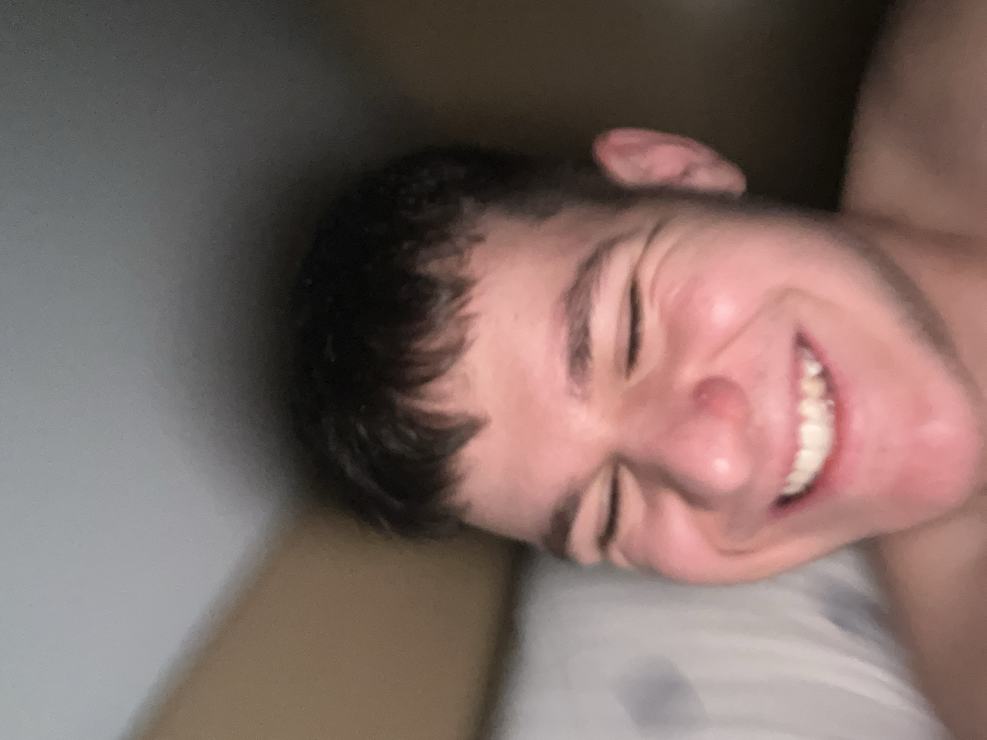 A close-up, angled portrait of a young man is presented indoors. He is lying down, with his head resting on a light-colored pillow. His eyes are closed, and he is smiling broadly, revealing his teeth. He has short, dark hair and fair skin. The background is a mix of light gray and darker brown tones. The location is not evident from the visual information provided. The city of Sofia, Bulgaria, cannot be confirmed based solely on the image.