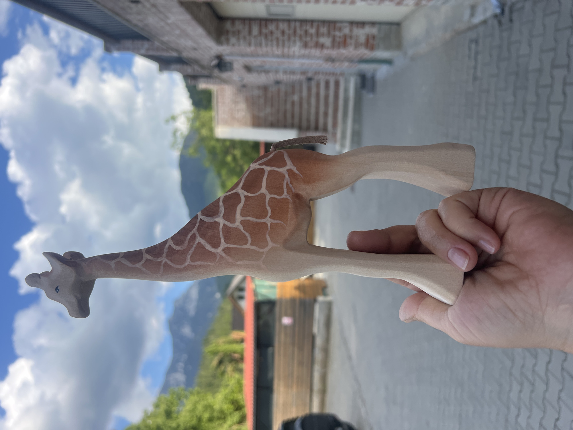A person's hand holds a wooden giraffe toy outdoors. The giraffe is light brown with darker brown spots. The background shows a paved area, a brick building with garages, trees, and mountains under a cloudy blue sky. The scene is brightly lit by daylight.