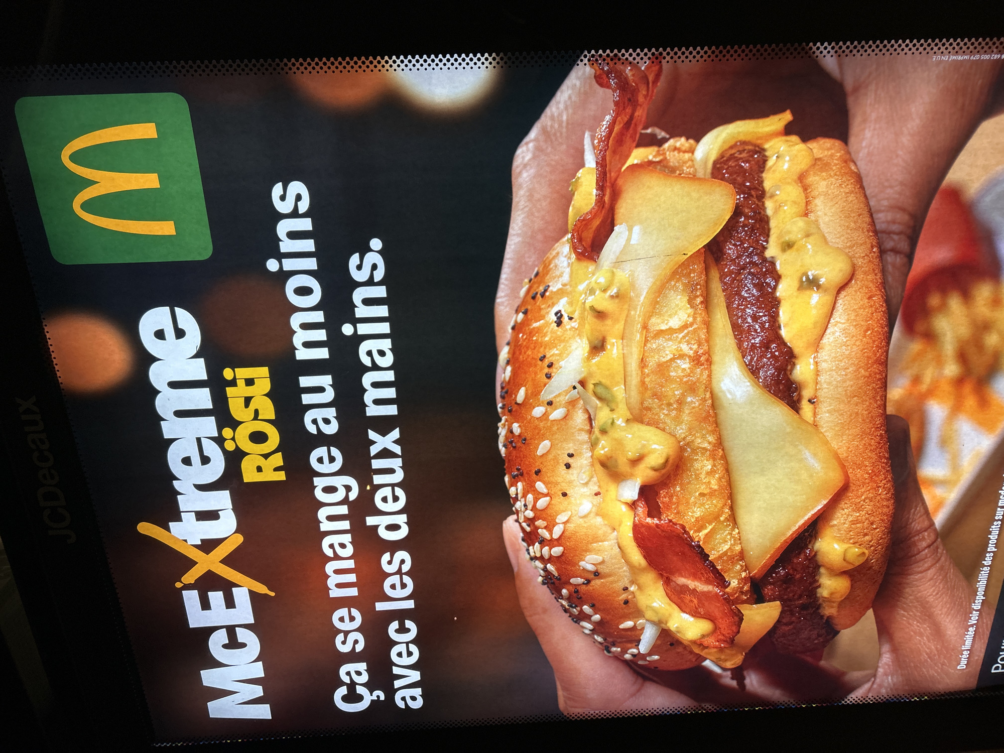 The image is a close-up photograph of an advertisement for McDonald's McExtreme Rösti, likely posted in Strasbourg, France. The left side of the image contains the McDonald's logo (a yellow "M" on a green background) and the product name, "McExtreme Rösti." Below the product name, in French, is the phrase "Ça se mange au moins avec les deux mains," meaning "It's eaten with at least two hands." The right side of the image is dominated by a close-up shot of the McExtreme Rösti sandwich. It shows a sesame seed bun holding what appears to be a beef patty, melted cheese slices, bacon, and some kind of sauce. The sandwich is being held by a person with dark skin. A tray with French fries can be seen slightly out of focus in the background. The advertisement is labeled at the bottom left corner as a JCDecaux ad. The image appears to be taken from inside a vehicle, evidenced by the windshield.