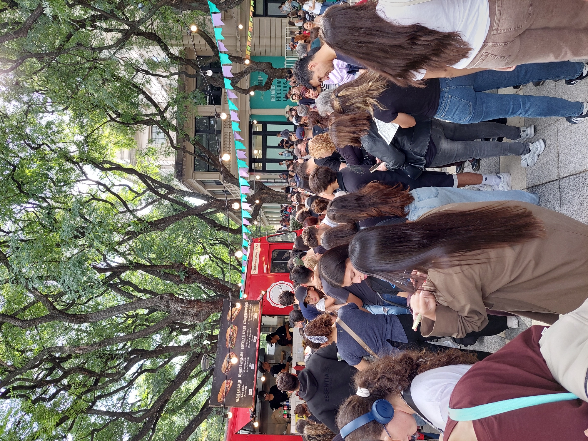The image shows a crowded street scene in Buenos Aires, Argentina. A red food truck is parked along the street, with a long line of people waiting to order food. The food truck menu visible in the image includes options such as "Vacio Ahumado", "Bondiola Ahumada", "Bondiola al Spiedo", and "Cordero Patagonico". Above the crowd, a string of colorful flags are hung between the trees, adorned with lights. The banner says "150 Anos Hipodromo Detodos". The trees cast shadows on the street and add to the vibrant atmosphere. Many people in the line have their attention focused on their phones, while others are engaged in conversations.