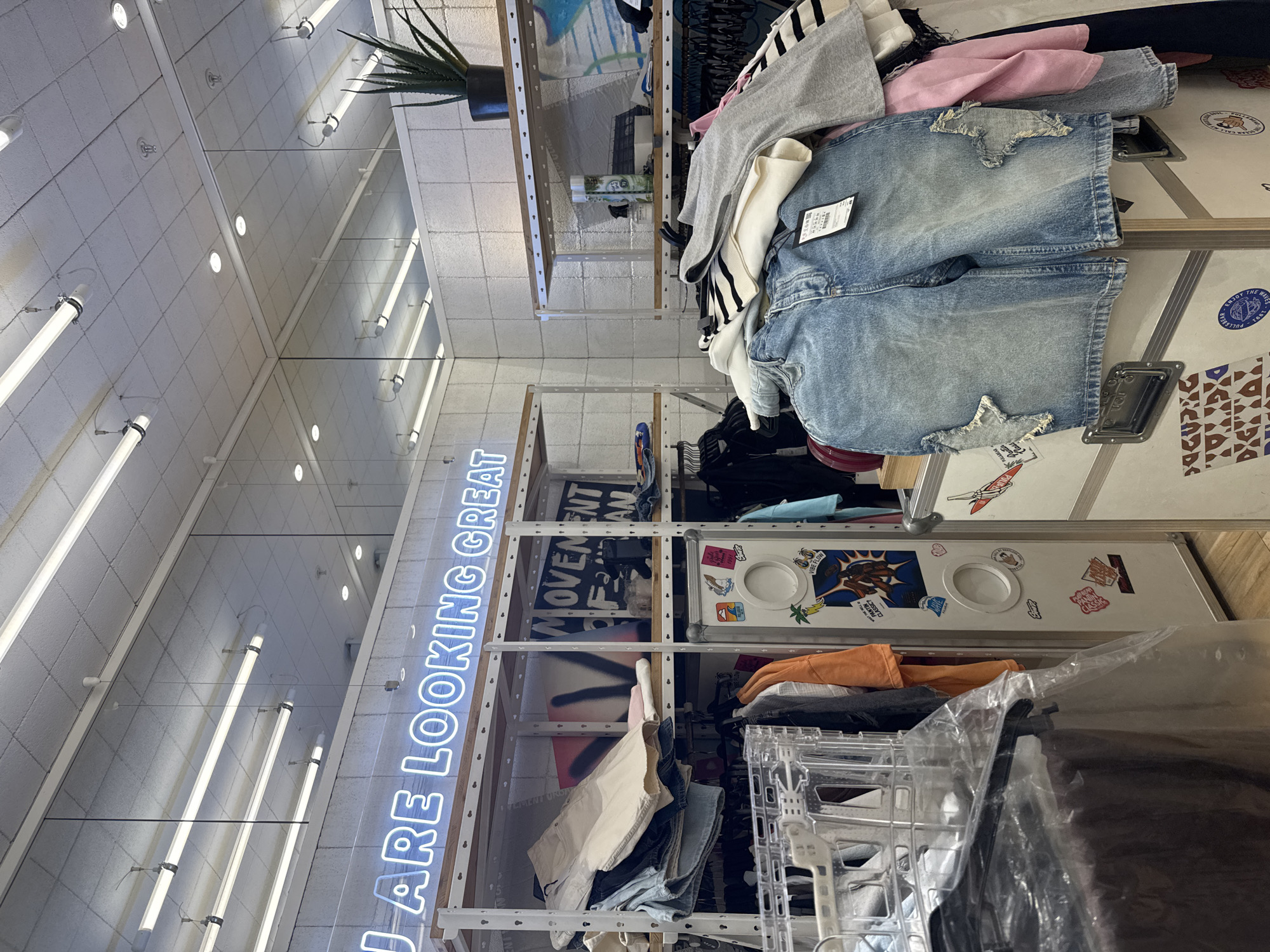 An indoor, slightly low-angle photograph captures a section of a clothing retail store. The location is an unknown city and country. No individuals are present in the frame.
The ceiling consists of white acoustic tiles, illuminated by multiple suspended fluorescent tube lights and recessed circular spotlights. Several sprinkler heads are also visible. A large, reflective mirror panel spans the upper section of the wall, reflecting the ceiling and its lighting, effectively extending the perception of the ceiling space.
The lower walls are surfaced with textured, light-colored square tiles. A prominent blue neon sign, horizontally mounted above a display system, reads "U ARE LOOKING GREAT".
Various items of clothing are displayed on white metal shelving units with light wood shelves, and stacked on large, white, metal-edged display cases covered in stickers. On the left and central shelving, folded garments, including light beige trousers and dark denim, are neatly stacked. Hanged clothing is visible beneath the shelves. The background of the shelving units features abstract art pieces or posters, one partially legible as "MOVEMENT OF MAN". A tall, narrow, white display unit with two circular cutouts is adorned with numerous stickers, including brand logos like "PULL&BEAR," and motifs such as palm trees, cartoon characters, and "VIBE FLOW."
In the foreground, a substantial pile of clothing rests on a white display case. This pile includes light-wash denim shorts featuring distinctive star-shaped distressed patches with raw edges on the thighs, a black price tag attached to them, along with folded striped sweaters, grey athletic wear, pink fabric, and white tops. A transparent plastic bag or garment cover partially obscures items in the bottom left.
On a shelf to the far right, a potted aloe vera plant, a green spray bottle, and other small accessories are visible. The flooring is a light-toned wooden surface. The store is brightly illuminated by artificial lighting, creating a clear and well-lit environment.