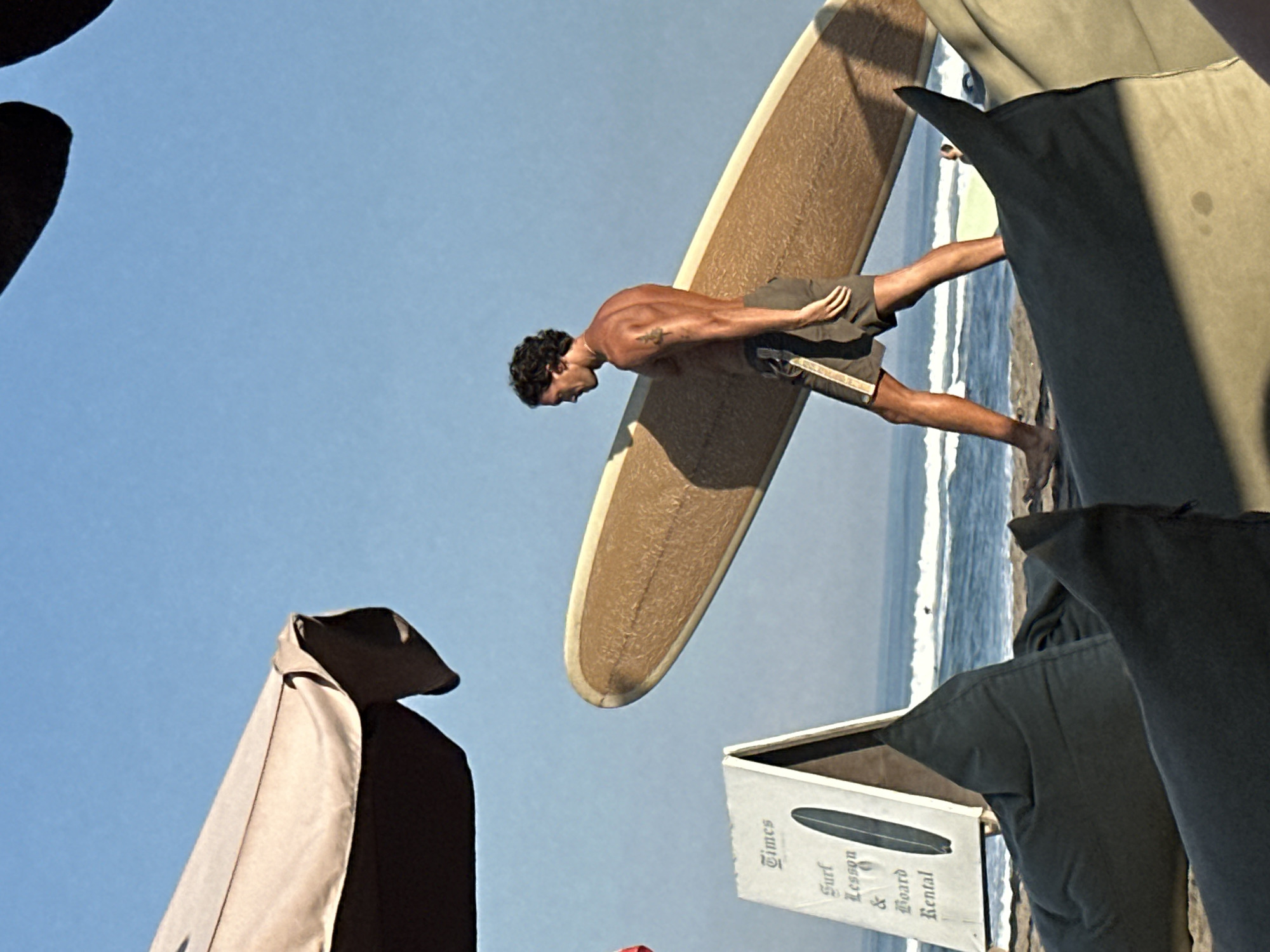 A shirtless man with curly dark hair walks across a sandy beach in North Kuta, Indonesia, carrying a light brown surfboard horizontally. He wears dark green shorts with a light stripe. In the background, the ocean has moderate waves, and a few surfers are visible. In the foreground, large, dark beanbag chairs are scattered on the sand. To the left, a sign advertising "Surf Lesson & Board Rental" stands propped up, and a portion of a light-colored beach umbrella is visible at the top left of the frame. The sky is clear and blue.