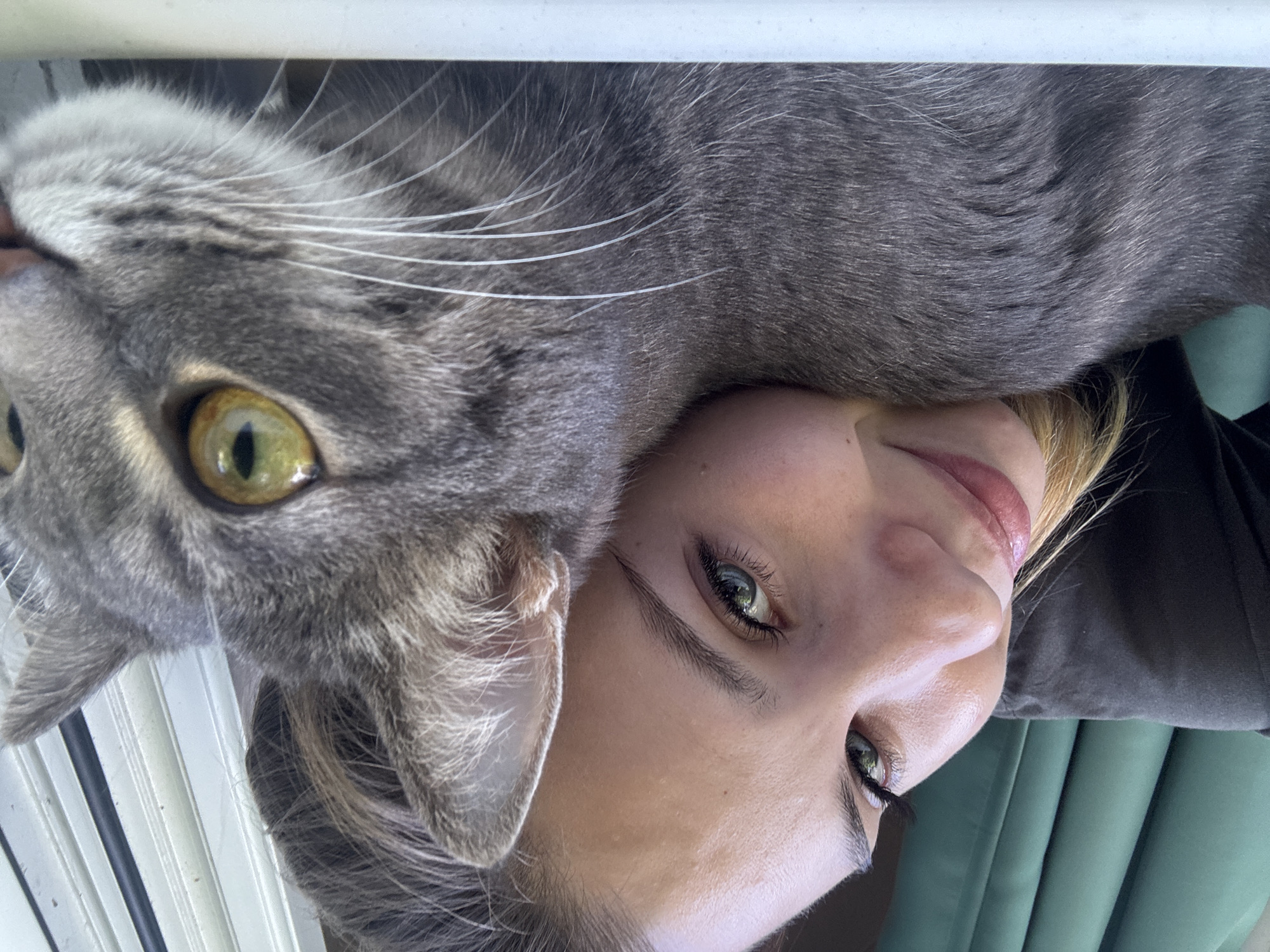 A close-up selfie shows a young woman with blonde hair and green eyes resting her head on a gray tabby cat. The woman is smiling, and her eyes are closed. The cat's eyes are open and looking at the camera. The background is blurred, but it appears to be an indoor setting with green curtains and a window with blinds. The image is taken from a high angle, with the camera looking down at the woman and cat. There is no information available regarding the location in the city of Chaikovsky, Russia.