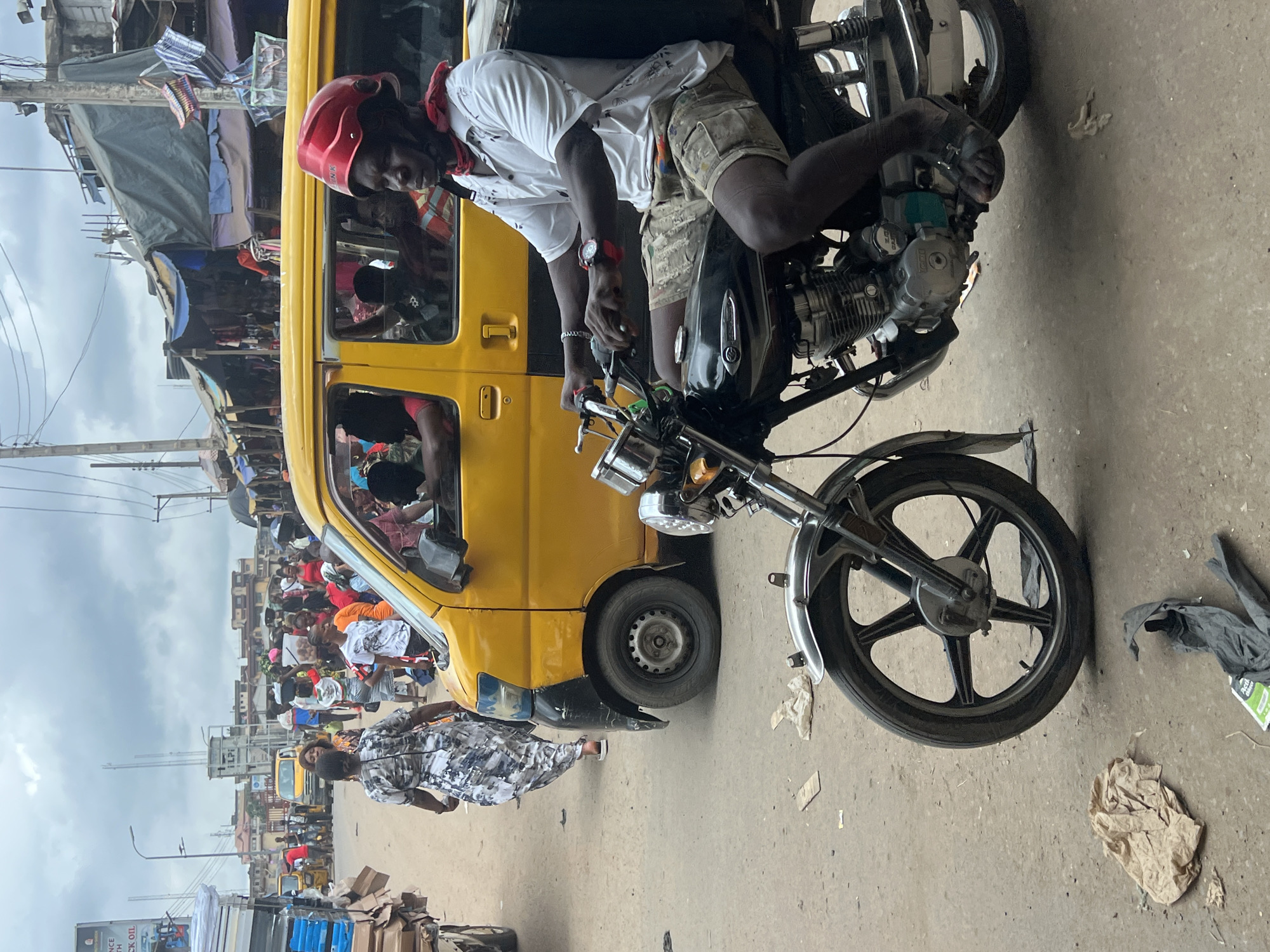 In Nkpor, Nigeria, a man wearing a red helmet and a white short-sleeved shirt with a tropical print is riding a motorcycle. He is wearing camouflage-patterned shorts and dark sandals. A red bandana is tied around his neck. He is looking to his right, towards a yellow mini-van. The mini-van has several passengers inside, visible through the windows. A woman in a patterned dress is walking away from the motorcycle and the mini-van. The scene appears to be a busy street, with market stalls and other vehicles visible in the background. The ground is unpaved and dusty, with some debris scattered on it. The sky is overcast.