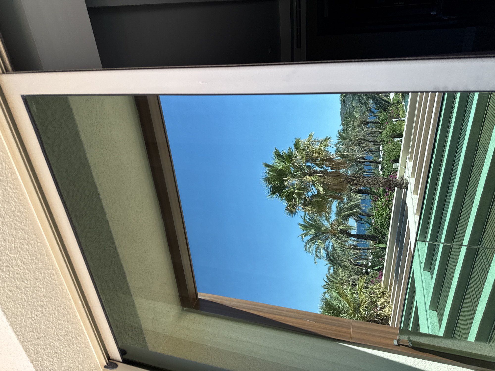 The image captures a view through a sliding glass door, likely in Bodrum, Turkey, looking out onto a resort-like setting. In the foreground, a patterned green surface with horizontal lines suggests a rooftop or a decorative element. Beyond this, a body of water, possibly a swimming pool, is visible, lined with white structures and lush greenery. A prominent cluster of palm trees stands tall against a clear blue sky, with more palm trees and other vegetation stretching towards the horizon. In the distance, rolling hills or mountains meet the sea, characteristic of the Bodrum coastline. The glass door frame is a light beige color, with a dark brown wooden trim around the opening. The interior of the room is dimly lit, contrasting with the bright outdoor scene. There are no people or animals visible in the image.