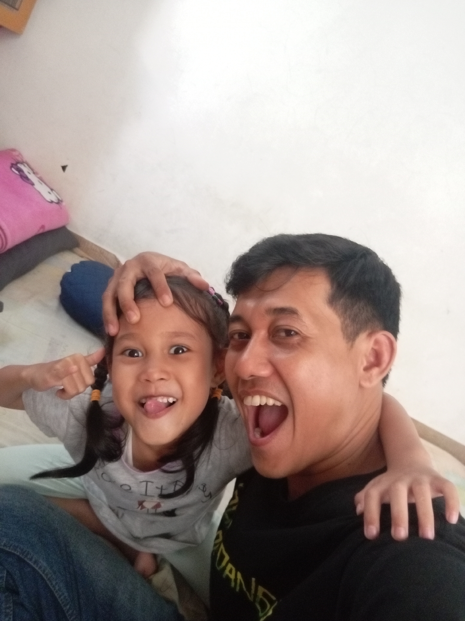 In Batam, Indonesia, a man and a young girl are taking a selfie. The man is in the foreground on the right, with his mouth open in a wide smile. He has dark hair and is wearing a black t-shirt. The girl is on the left, with her tongue sticking out and her index finger touching her forehead, as if mimicking a playful gesture. She has dark hair in pigtails and is wearing a gray t-shirt. The man's arm is around the girl's shoulders, and his hand is resting on her head. In the background, there is a white wall and some soft furnishings, including a pink pillow with a cartoon character on it and a blue bolster pillow. The overall impression is one of a warm and affectionate moment captured between a father and daughter.