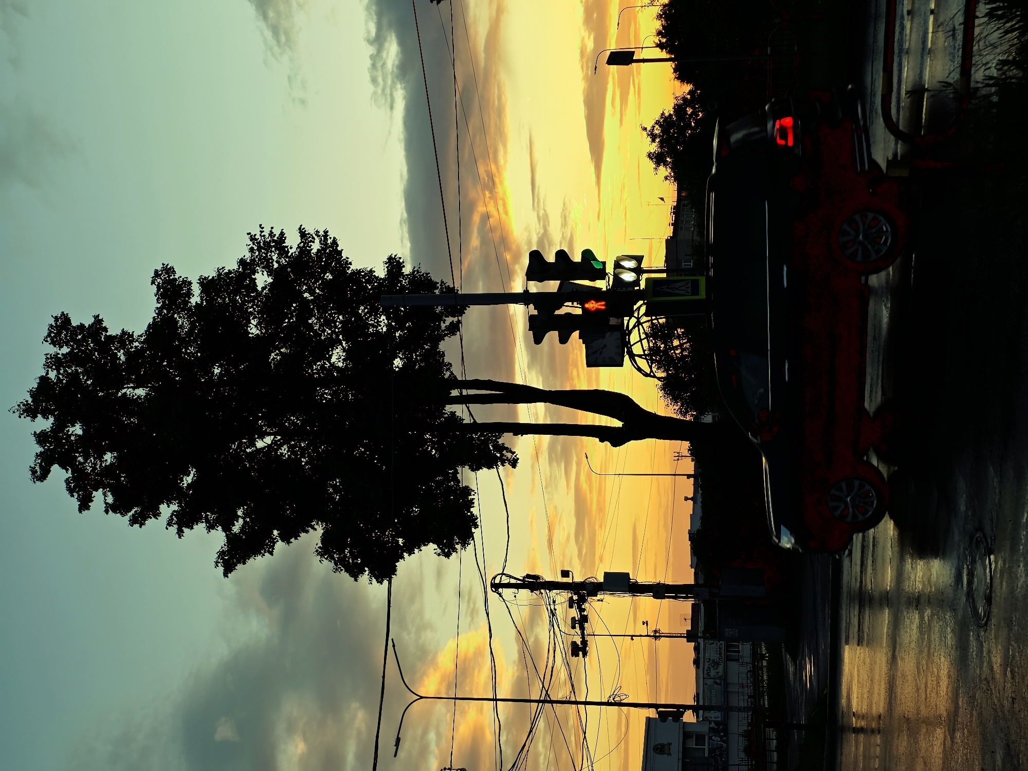 The image depicts a street scene at dusk in Chaikovsky, Russia. A large tree with dark foliage stands silhouetted against a vibrant sunset sky with hues of yellow, orange, and teal. Power lines and streetlights are visible, along with a traffic light showing a red pedestrian signal and a green vehicle signal. A dark-colored SUV, partially obscured by its silhouette and with visible red accents, drives on the wet asphalt road, reflecting the sky's colors. The road surface appears to be a typical urban asphalt, with some puddles and a circular manhole cover visible. Buildings and trees are seen in the background. The overall atmosphere is one of twilight in a city setting.