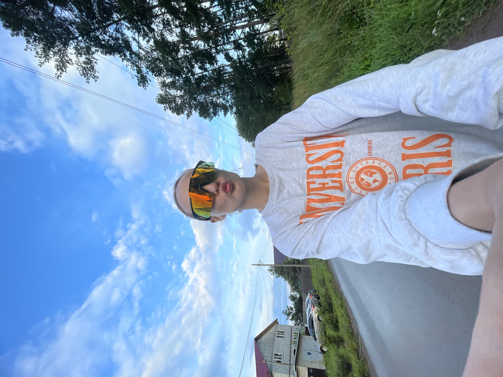 A person wearing large, reflective, orange-tinted sunglasses and a light gray sweatshirt with orange lettering that reads "UNIVERSITY OF SCIENCE GROWING FROM PARIS" is holding a camera to take a selfie outdoors. They are outdoors on a paved road. To the left of the road, there is a two-story house with a gray facade and a burgundy roof, and a few cars are parked on a patch of grass. To the right of the road, there are tall green trees and thick grass. The sky is blue with scattered white clouds. The image appears to be taken in or near the village of Podshivalovo, Russia.