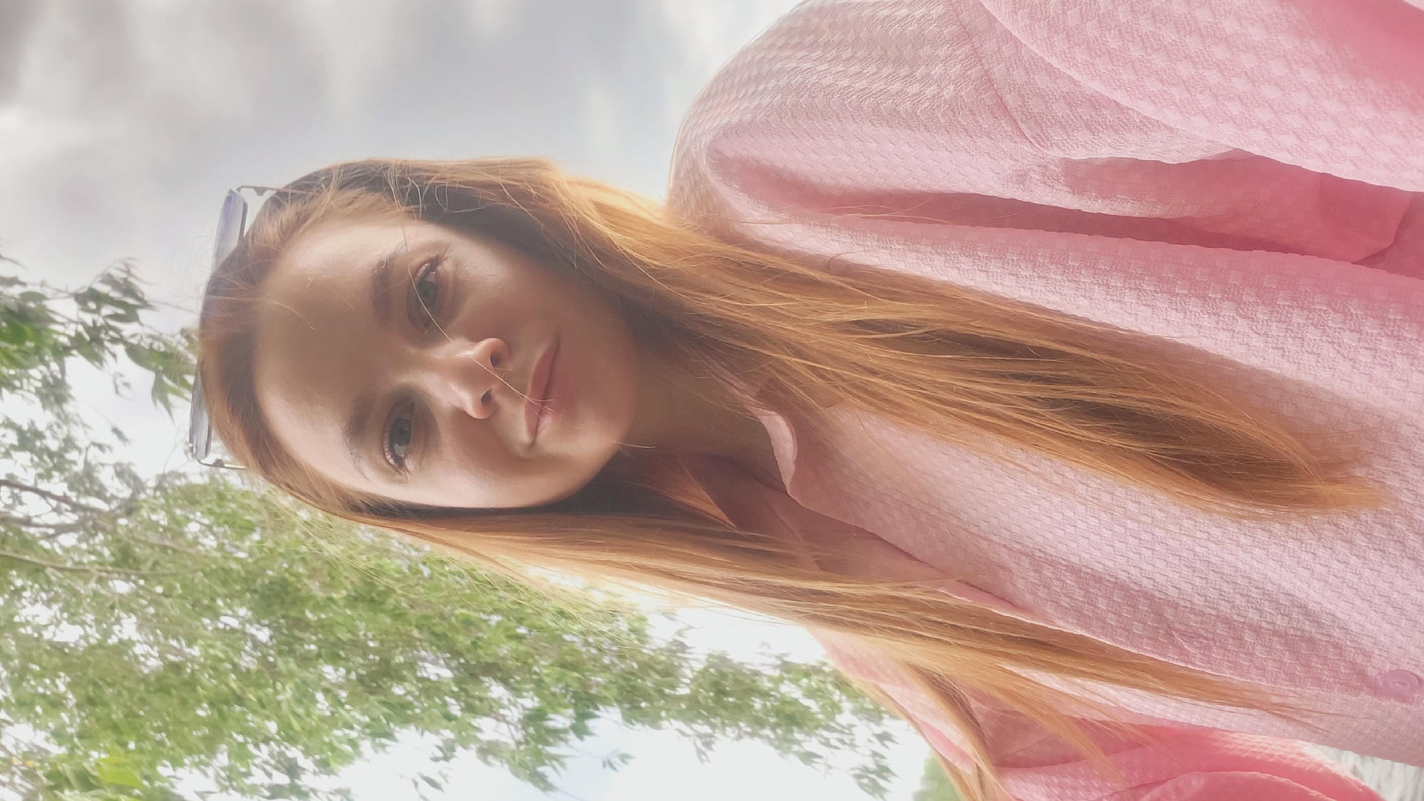 A low-angle, selfie-style photograph features a woman with reddish-brown hair and pale skin wearing a pink, textured button-down shirt. She is positioned slightly off-center and tilted to the right. Sunglasses are perched on her head. In the background, blurred green foliage suggests a tree or trees, while a pale, overcast sky fills the upper portion of the frame. There is no visible indication of the location being Chaikovsky, Russia.