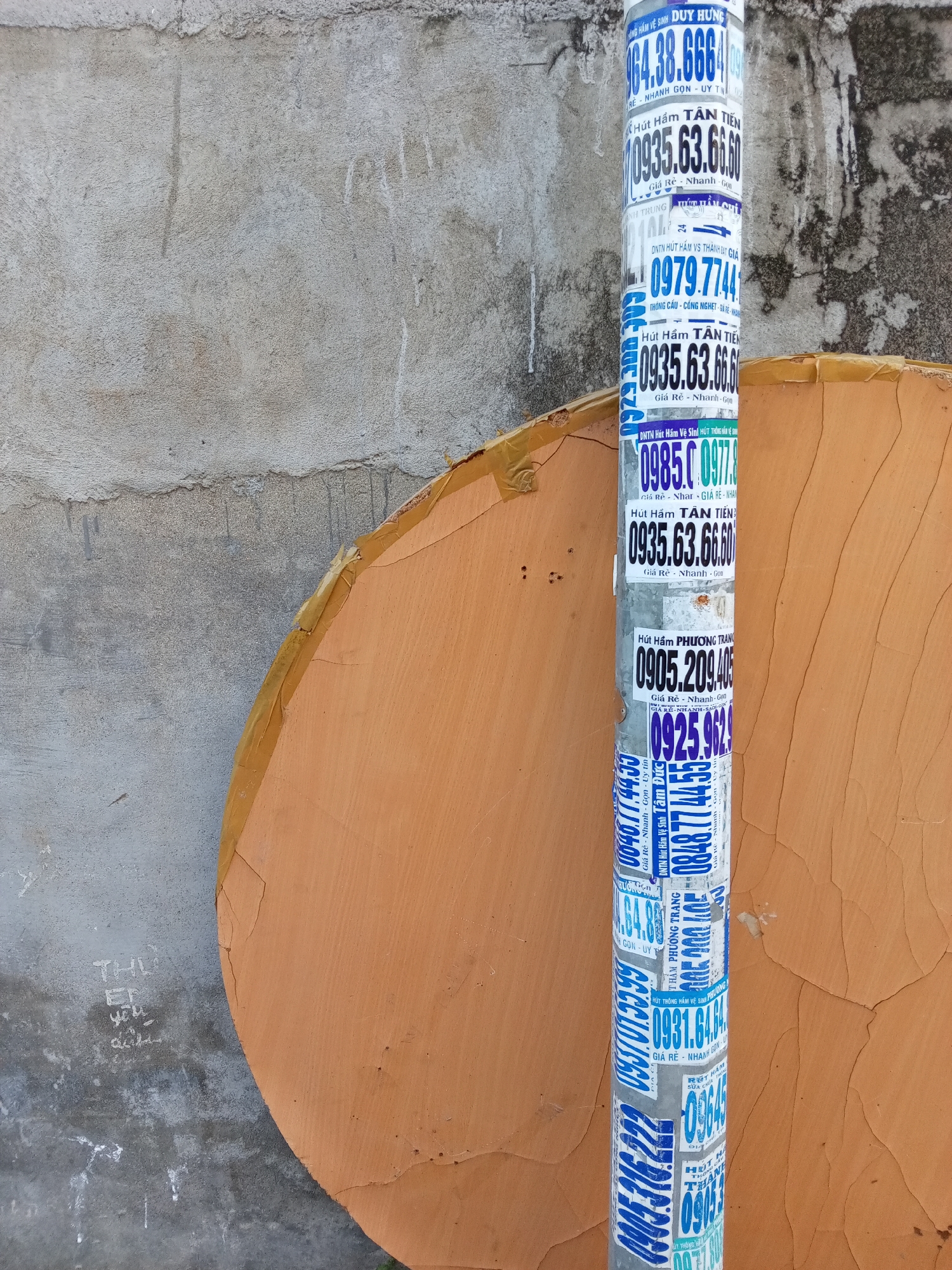 The image shows a utility pole in an urban area in Nha Trang, Vietnam. The pole is covered in advertisements for septic tank cleaning services, each displaying a phone number and the phrase "Giá Rẻ - Nhanh - Gọn" (Cheap - Fast - Convenient). A weathered, orange-colored circular object is behind the pole. A concrete wall is to the left of the pole. There is graffiti on the wall.
