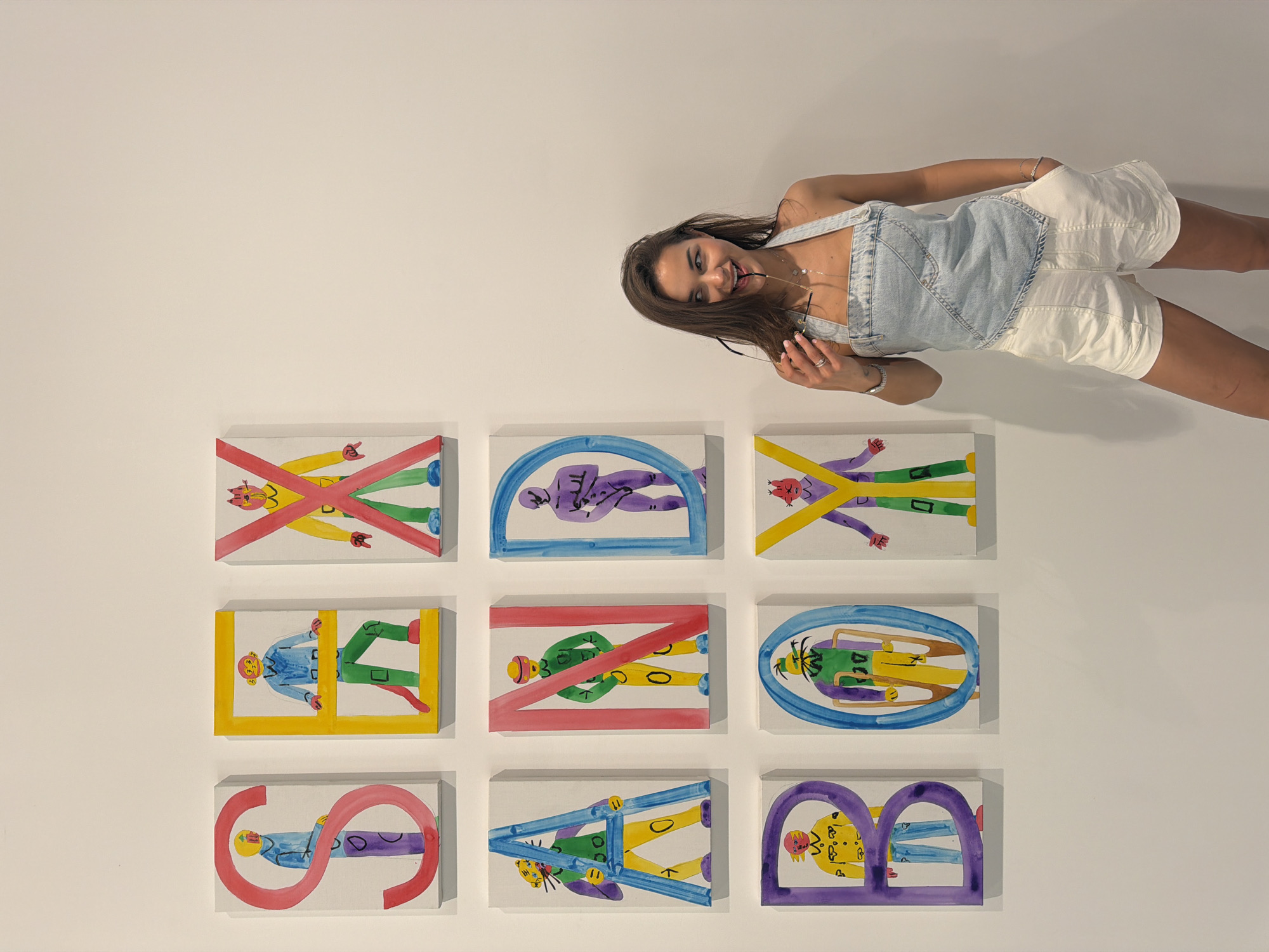 A young woman in a light blue denim top and white shorts is posing next to a display of nine small, rectangular canvases. The canvases are arranged in a 3x3 grid on a white wall. Each canvas features a cartoon-style drawing of a person outlined by a colorful geometric shape. The shapes include an "X," a half-circle, a straight line, a square, an oval, and a figure-eight. The woman is angled in the frame, with her head and upper torso positioned towards the top right corner and her legs stretching towards the top right edge. She is smiling and looking at the camera. The background is a plain white wall. The location in Chaoyang District, China, is not discernible from the image.