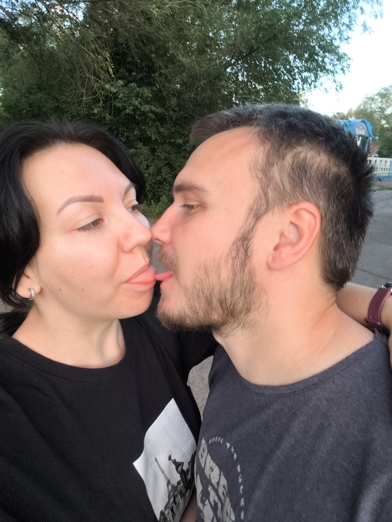 In Orenburg, Russia, a close-up, outdoor photograph captures a couple engaging in a playful, intimate moment. The woman, on the left, has dark hair and is wearing a black t-shirt with a white graphic of the Eiffel Tower. Her right ear is adorned with a silver hoop earring. The man, on the right, has short, dark hair and a light beard, and is wearing a dark grey t-shirt. Both individuals are leaning towards each other, with their faces very close. The man's tongue is extended, touching the woman's tongue. They appear to be in a park or an outdoor recreational area, as suggested by the blurred background of green foliage and trees, and what appears to be a distant amusement park ride with the word "GRAND" visible. The lighting suggests it is late afternoon or early evening.