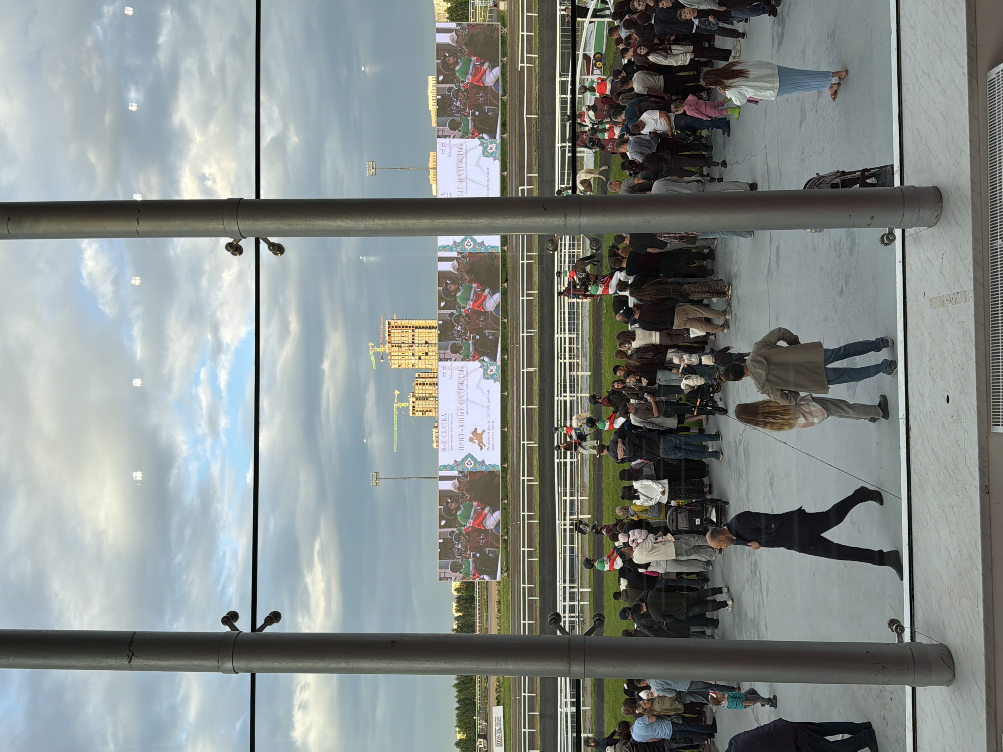 This is a photograph of a horse race event, likely a flat race, as indicated by the racetrack visible in the background and the large screen displaying images of horses and riders. The image is a reflection, possibly in a glass building or a large window, which creates a mirror image of the scene.
The subjects in the image are a crowd of people gathered to watch the race, and the racetrack itself. The people are standing and appear to be looking towards the racetrack, suggesting they are spectators. Some individuals are dressed casually, while others are in more formal attire. The screen shows a close-up of horses and jockeys, with text in Russian. The text on the screen reads "8-Я СКАЧКА" (8th Race), "Дистанция 500 метров" (Distance 500 meters), "ПРИЗ «ЮНЫЕ НАДЕЖДЫ»" (Prize "Young Hopes"), and "Скачка на пони" (Pony Race). There is also a mention of a prize fund of 100,000 rubles.
The scene is set outdoors, with a racetrack visible. The sky in the reflection appears cloudy, suggesting it might be an overcast day or late afternoon. The overall atmosphere seems to be one of anticipation and enjoyment of the sporting event. The reflection adds a unique perspective, showing the sky and buildings above the actual event. The presence of multiple screens and a grandstand (implied by the crowd's position) suggests a well-established venue for horse racing.