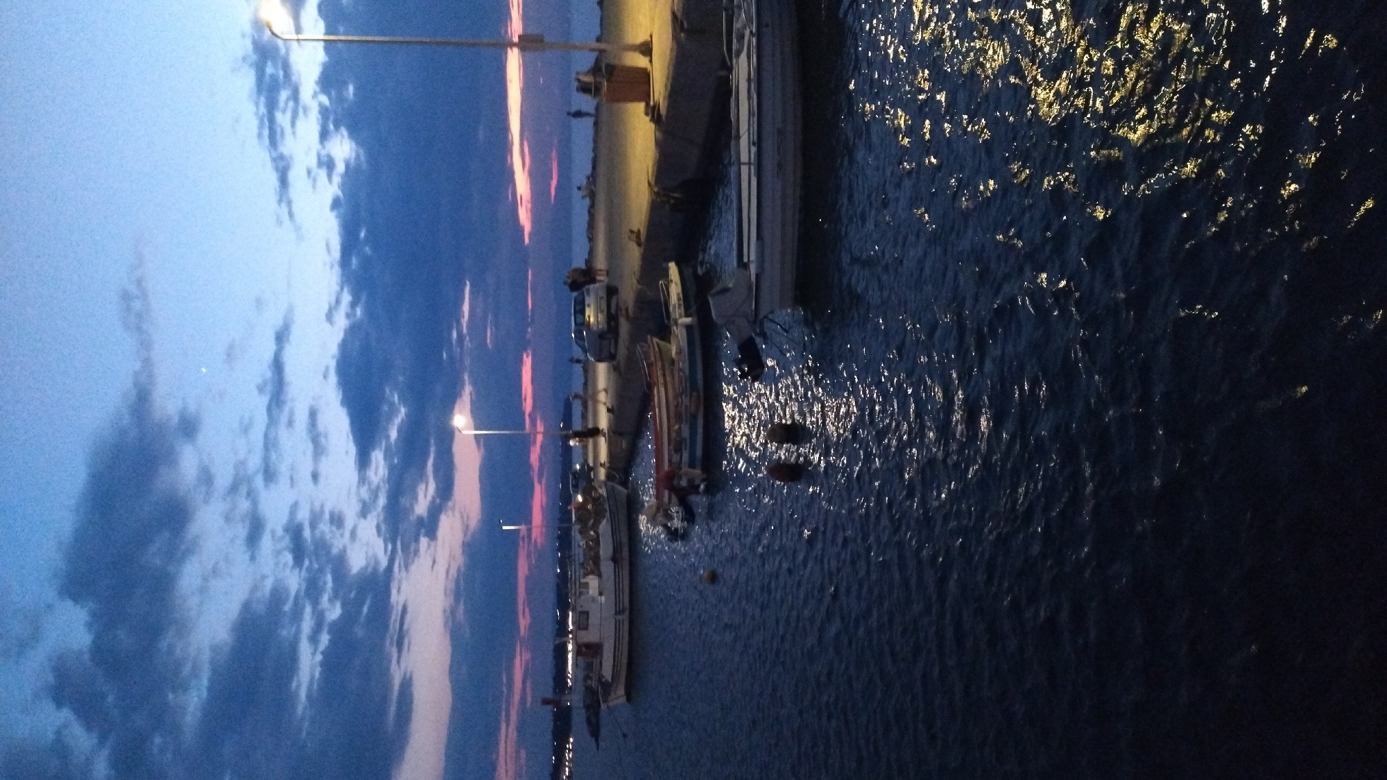 The image depicts a twilight scene at a harbor in Neoi Epivates, Greece. The sky is a gradient of deep blue with streaks of pink and purple near the horizon, illuminated by scattered dark clouds and the setting sun. Several boats are moored in the calm, dark blue water, reflecting the lights from the shore. The foreground shows the dark water with highlights from artificial lights. A concrete pier runs along the right side of the frame, illuminated by streetlights. On the pier, a few people are visible, some standing and others walking. A silver car is parked on the pier, and a trash receptacle is located near the edge. In the background, the coastline is lined with lights, suggesting the presence of a town or village. A lighthouse or a tall structure is faintly visible on the left horizon. The overall atmosphere is serene and coastal.