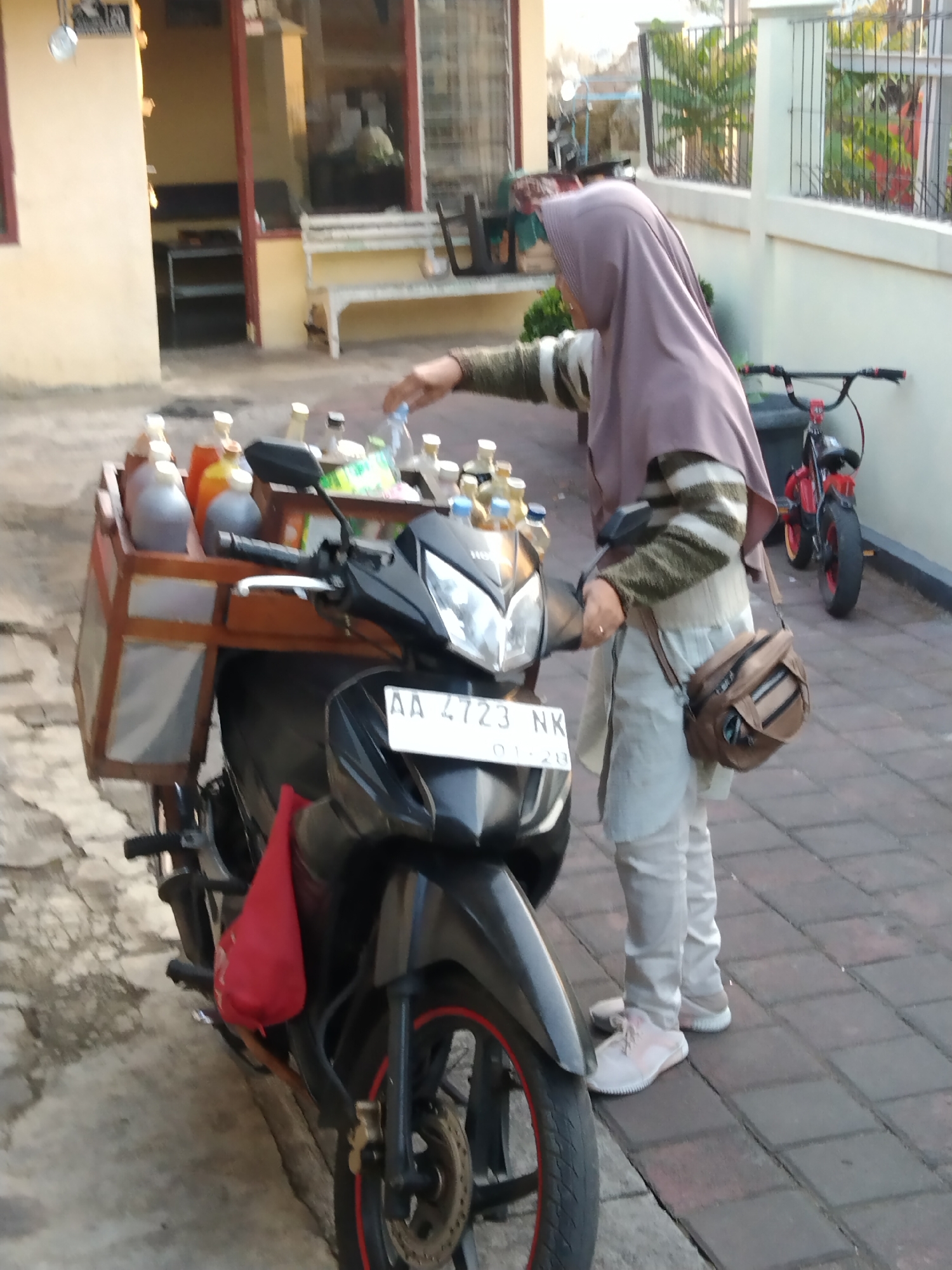 In Magelang, Indonesia, a woman wearing a purple hijab and a striped long-sleeved shirt is standing next to a black motorcycle. The motorcycle is equipped with a wooden crate on the back, containing multiple bottles filled with liquids of various colors, suggesting it is used for selling drinks. The woman's right hand is reaching towards one of the bottles. She is wearing light-colored pants and pink sneakers. A brown crossbody bag is slung over her shoulder. In the background, a red bicycle is leaning against a white fence. The scene appears to be an outdoor setting, possibly a street or sidewalk, with patterned brick flooring. A building with yellow walls and a red-trimmed doorway is visible on the left side of the image.