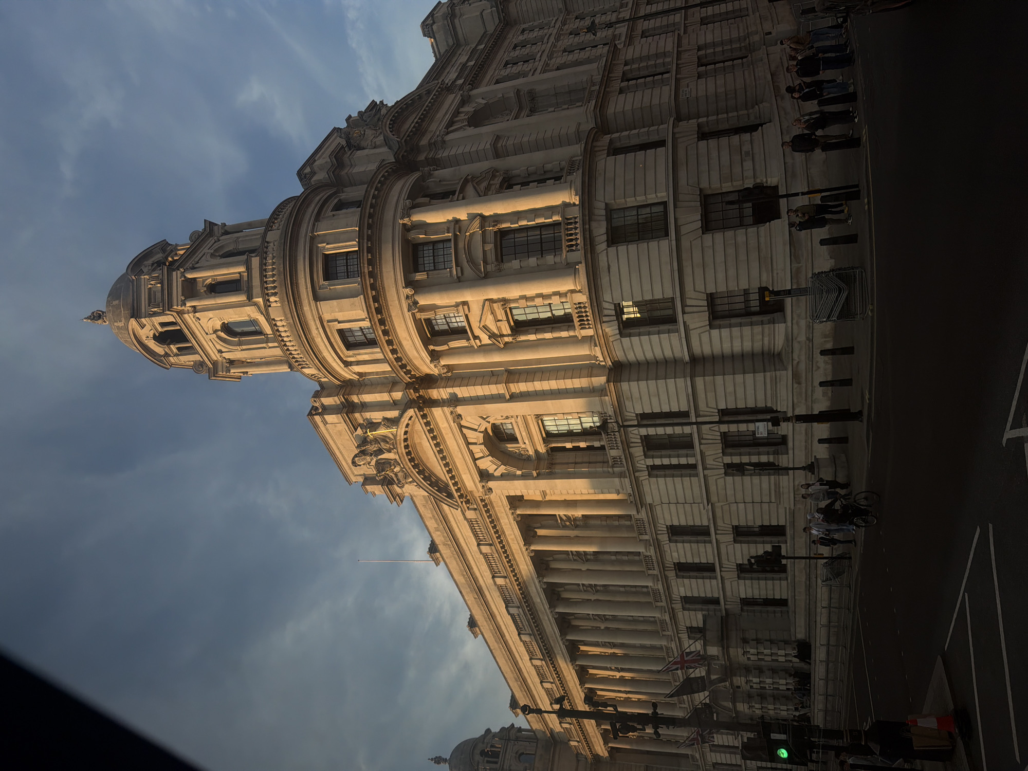 The image depicts a building in London, United Kingdom, likely Whitehall, photographed at an oblique angle. The building features classical architecture with ornate details, including columns, cornices, and a domed structure. A line of people are standing along the building's side facing the street. A street runs along the building and there are traffic lights and bollards visible. The sky is cloudy and the lighting suggests either sunrise or sunset.