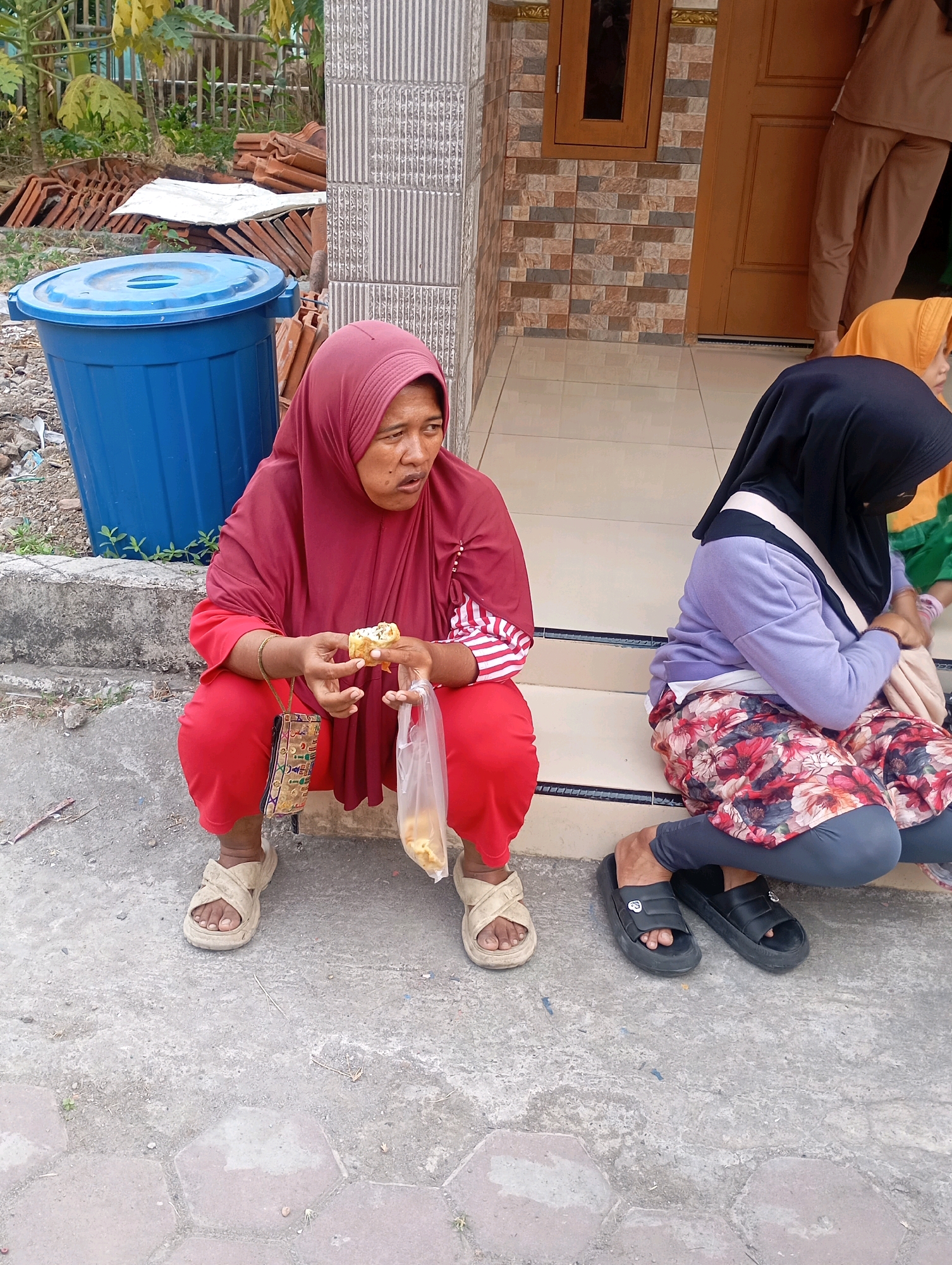 In Babelan, Indonesia, two women are seen sitting on a concrete walkway in front of a building. The woman on the left, wearing a maroon hijab and red pants, is squatting and holding a piece of food in her hand. She also holds a clear plastic bag containing food. She wears light-colored sandals. To her right, a second woman, wearing a black hijab, a purple top, and a floral-patterned skirt over grey leggings, is also squatting. She wears black slip-on sandals. The environment includes a blue trash bin, stacks of terracotta roof tiles, and vegetation in the background. The building features tiled walls and a brown door.