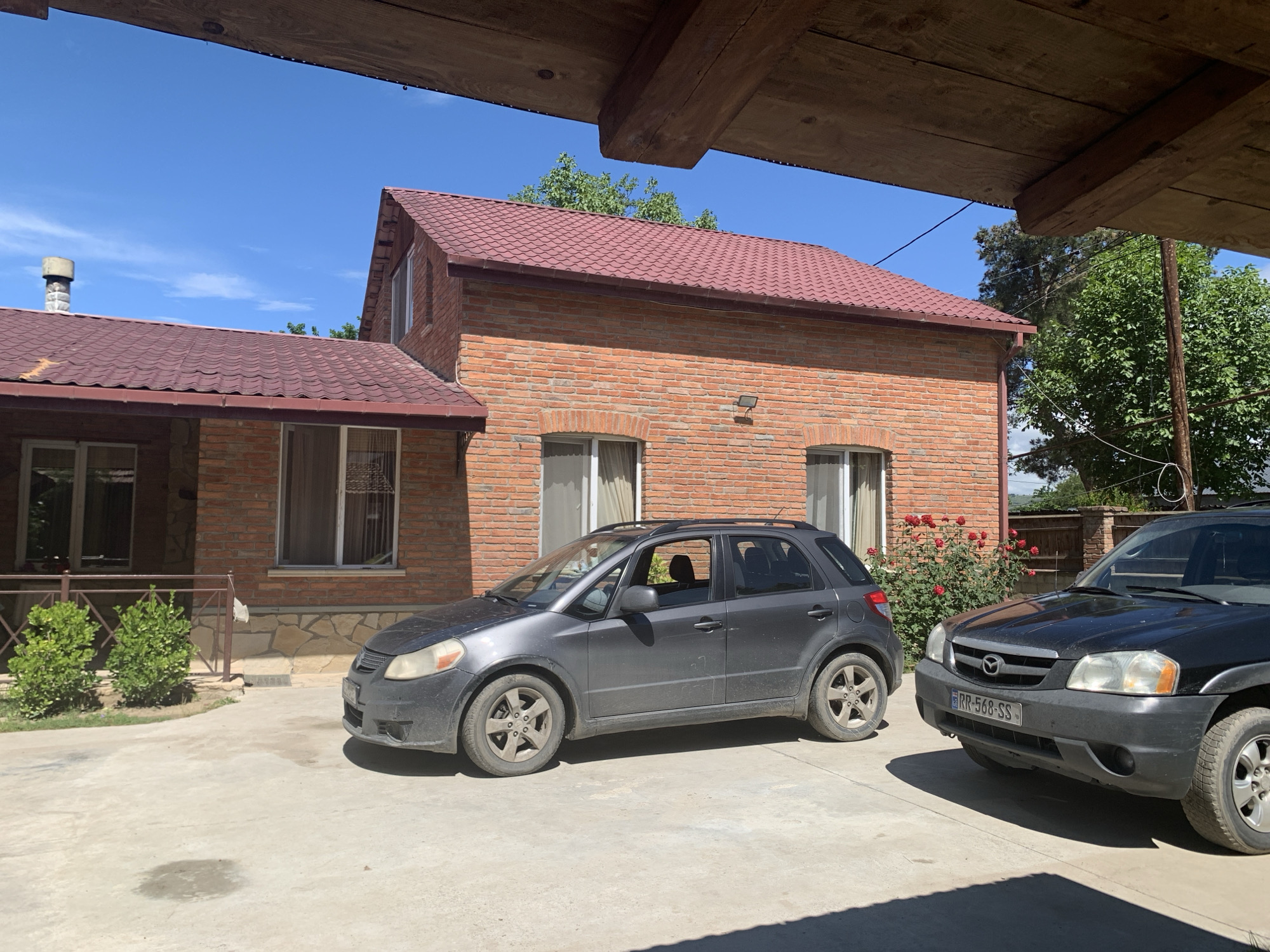 A dark gray Suzuki SX4 hatchback is parked on a concrete driveway in front of a red brick house with a burgundy metal roof. The house has two stories and several windows. To the right of the Suzuki, a black Mazda Tribute SUV is also parked on the driveway. Red roses are blooming in a bush to the right of the house. The sky is clear blue. The image is taken from a low angle, looking up at the house and cars, with a wooden overhang in the foreground. The scene appears to be a residential area.