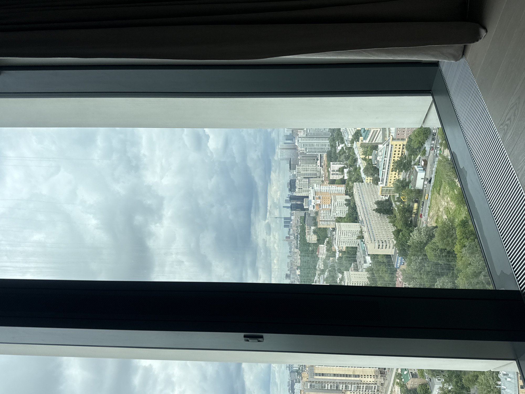 The image displays a high-angle view of a cityscape as seen through a large window, which is rotated 90 degrees clockwise from its correct orientation.
In the immediate foreground, the dark frame of the window is prominent, dividing the scene horizontally. A thin white sill or frame component runs parallel to the dark frame. Along the upper right edge of the image (which would be the top right if rotated correctly), a portion of what appears to be a sofa or upholstered furniture is visible. Below the window frame on the right side, there's a section of a white, slatted radiator or vent running along a light wooden floor, indicating an indoor setting.
Through the window, an expansive urban landscape stretches into the distance under an overcast sky. Numerous multi-story buildings, varying in height and architectural style, fill the scene. Many appear to be residential apartment blocks, characterized by their uniform, blocky designs. Interspersed among the buildings are significant areas of greenery, including trees and parks, suggesting a mix of built-up and natural environments. Roads with a few visible vehicles wind through the city. The sky is dominated by thick, grey clouds, indicating an overcast day.
Based on the provided context, the location is Khimki, Russia. No specific activity is depicted, only the static view of a city. There is no visible text in the image.