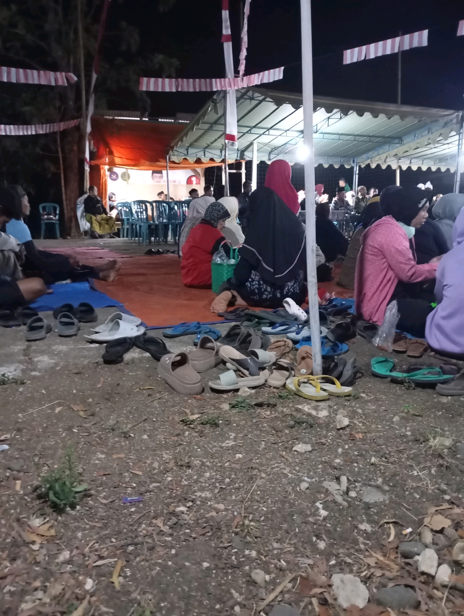 The image depicts an outdoor gathering at night in Wonogondo, Indonesia. A group of people, some wearing headscarves, are seated on the ground on a red mat and on chairs under a tent. Many pairs of footwear, including sandals and flip-flops, are scattered on the ground in the foreground. The scene is illuminated by artificial lighting, and decorative banners are visible in the background. The ground is covered in dirt and debris.