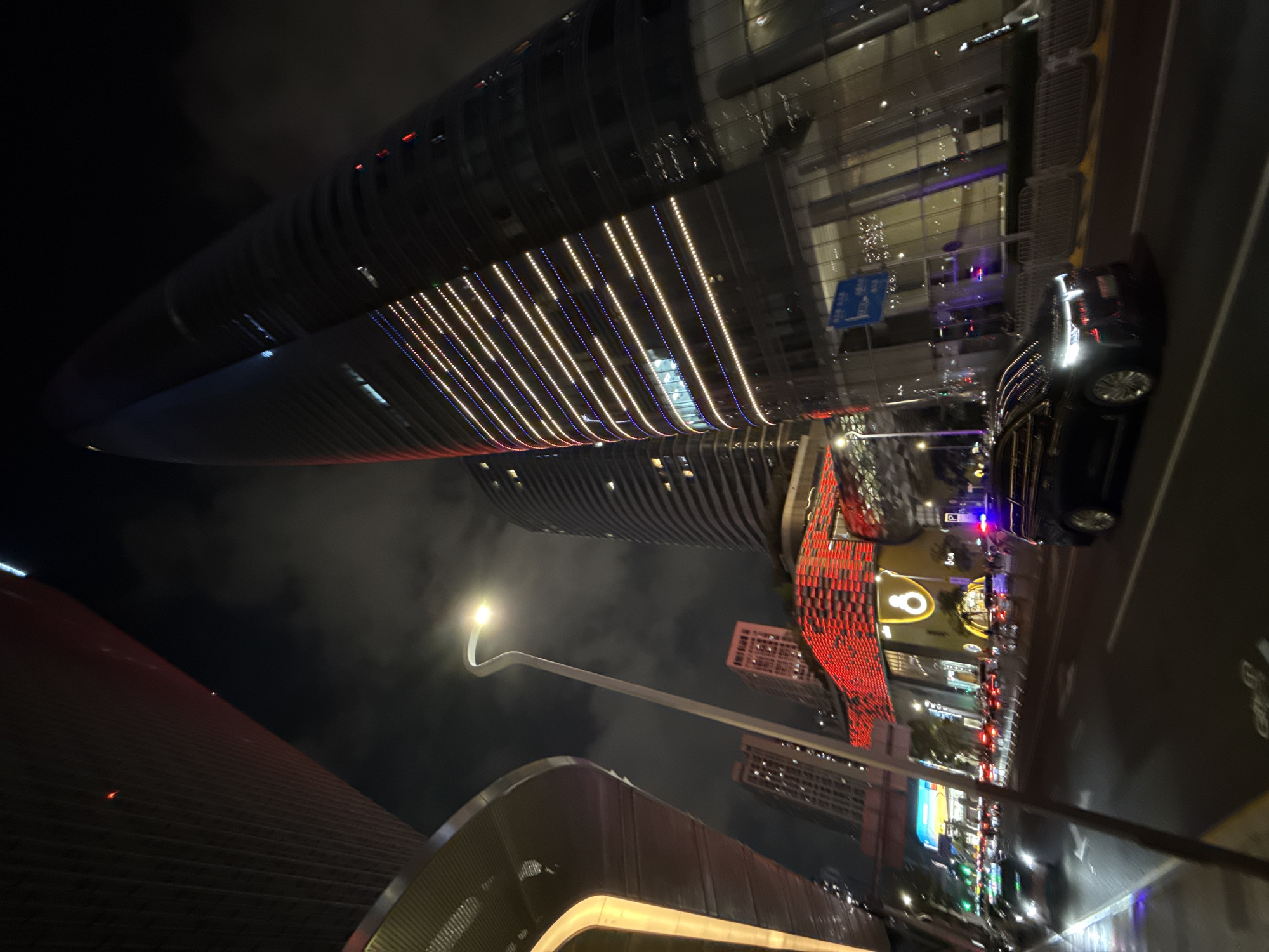 The image captures a night scene in Futian District, Shenzhen, China, characterized by modern architecture and urban illumination. Towering skyscrapers dominate the upper portion of the frame, featuring illuminated linear patterns and reflective surfaces. A sleek black car is parked on the roadside, its headlights contributing to the ambient light. A distinctive street lamp with a curved design stands out, providing localized lighting. Further into the scene, buildings with red and yellow color schemes add visual interest. Overall, the photograph depicts an urban landscape at night, showcasing the architectural design and lighting common in Futian District.