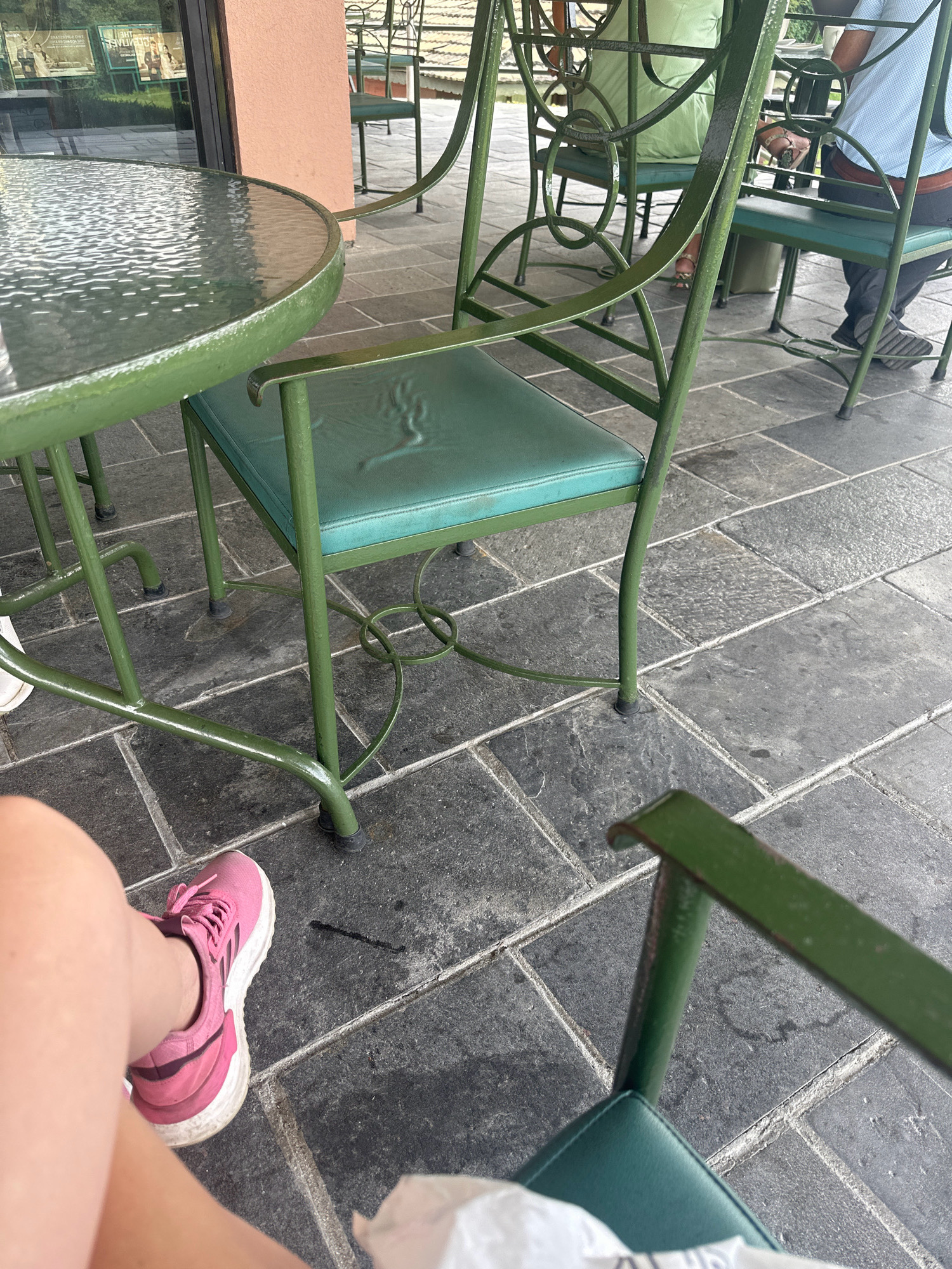 An outdoor seating area with multiple green metal chairs and a circular glass-top table is pictured on a grey tiled patio in Kageshwori Manohara, Nepal. A person's legs and feet, wearing pink athletic shoes, are in the foreground. In the background, a person in a light blue shirt and dark pants is seated, and another person wearing a light green garment is also seated. Behind the chairs, a building with a pink facade and large windows is visible, and beyond that, foliage and a glimpse of a pond or water feature can be seen. The scene appears to be in a restaurant or cafe setting.