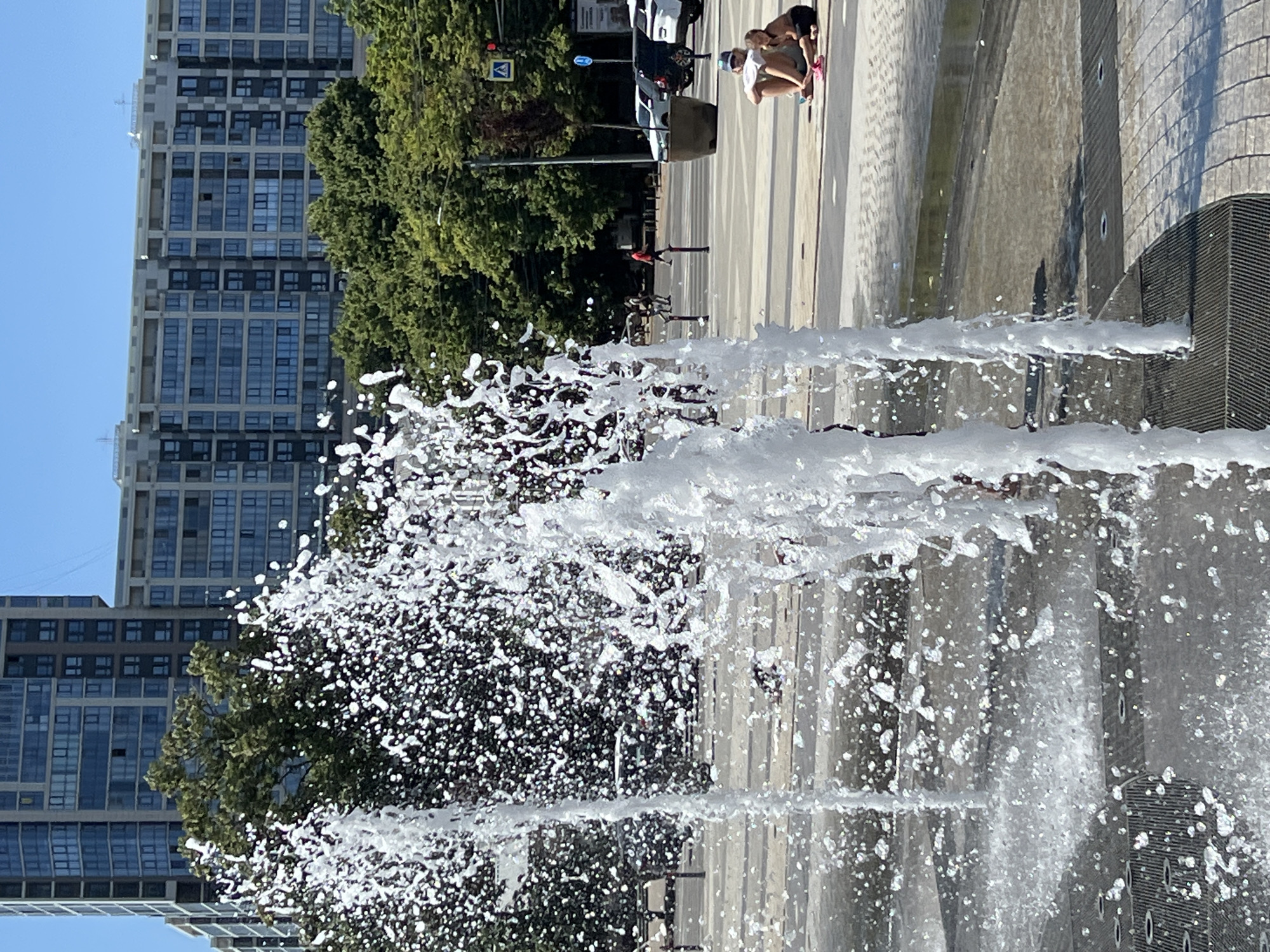 In Krasnodar, Russia, a public fountain is operating, with water spraying upwards in the foreground. Behind the fountain, a modern apartment building with numerous windows dominates the background, indicative of urban development. The sky is clear and blue, suggesting a sunny day. Two individuals, a man and a child, are seated on the ground near the fountain, appearing relaxed. In the middle ground, cars are parked on a paved surface, and a pedestrian crossing is visible. Trees line the street, providing a touch of greenery against the urban backdrop. The scene captures a moment of leisure in a city environment, with the fountain providing a refreshing element on a warm day.