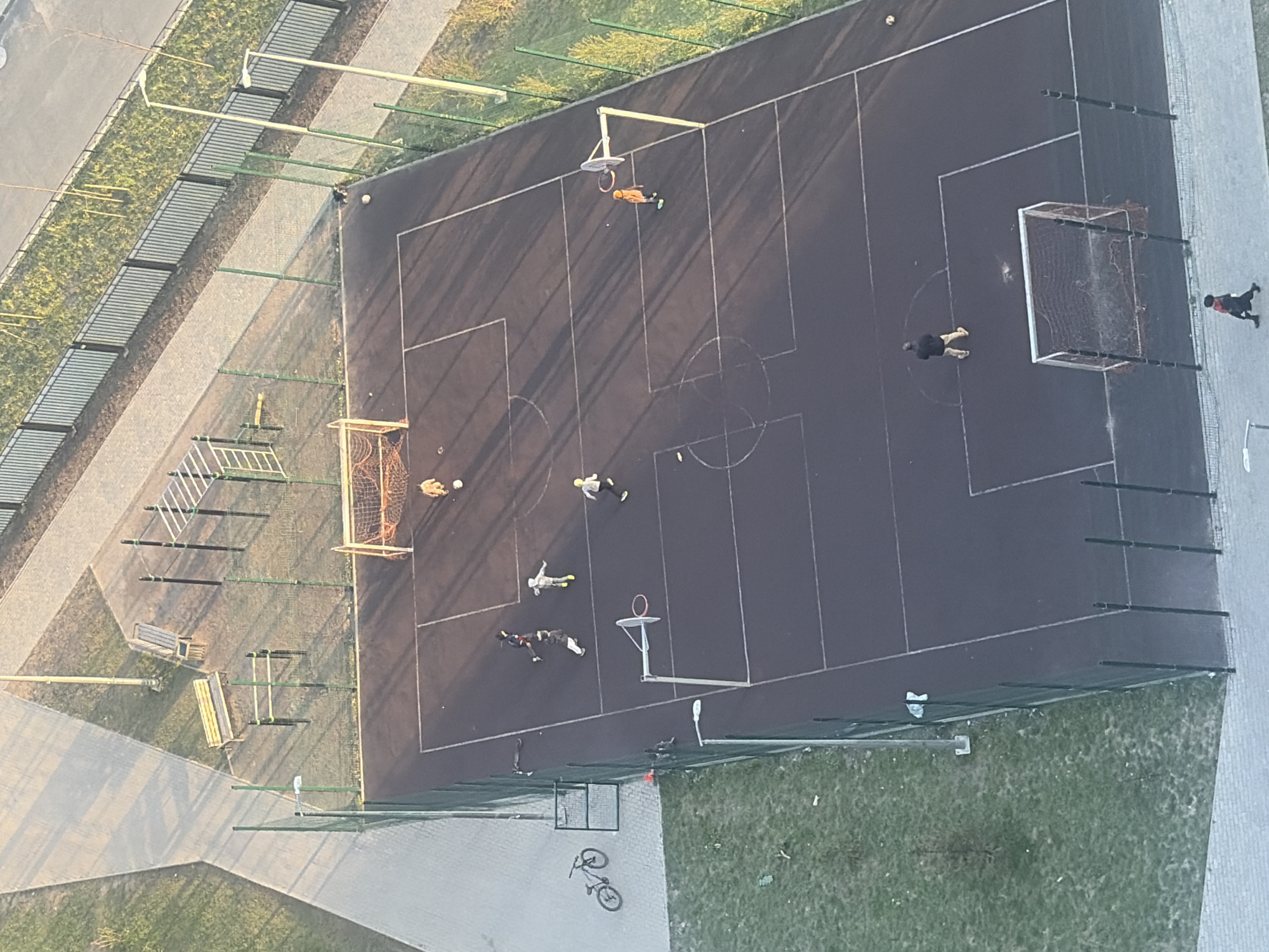 This aerial view depicts a multi-sport court in Saint Petersburg, Russia, likely during the late afternoon or early morning, indicated by long shadows stretching across the playing surface. The court features a dark grey or black asphalt surface marked with white lines for both soccer and basketball. It is enclosed by a green mesh fence on at least two sides.
Approximately eight to ten people, appearing to be children or teenagers, are present on and around the court. Several individuals are actively engaged in play: two people are near the left soccer goal with a ball, and another is near the basketball hoop. Other individuals are standing, walking along the paved perimeter, or lying down on the court. One person in a black jacket and light pants is near the right soccer goal, while another in a red and black outfit walks on the paved path adjacent to the court.
Key objects include two soccer goals with nets (one orange, one white), a basketball hoop, and at least two visible balls. Outside the court, to the left, there's exercise equipment with parallel bars and benches. A bicycle is lying on the ground near the bottom right corner, just outside the fenced area. The court is surrounded by paved walkways and grassy patches. No discernible text is visible in the image.