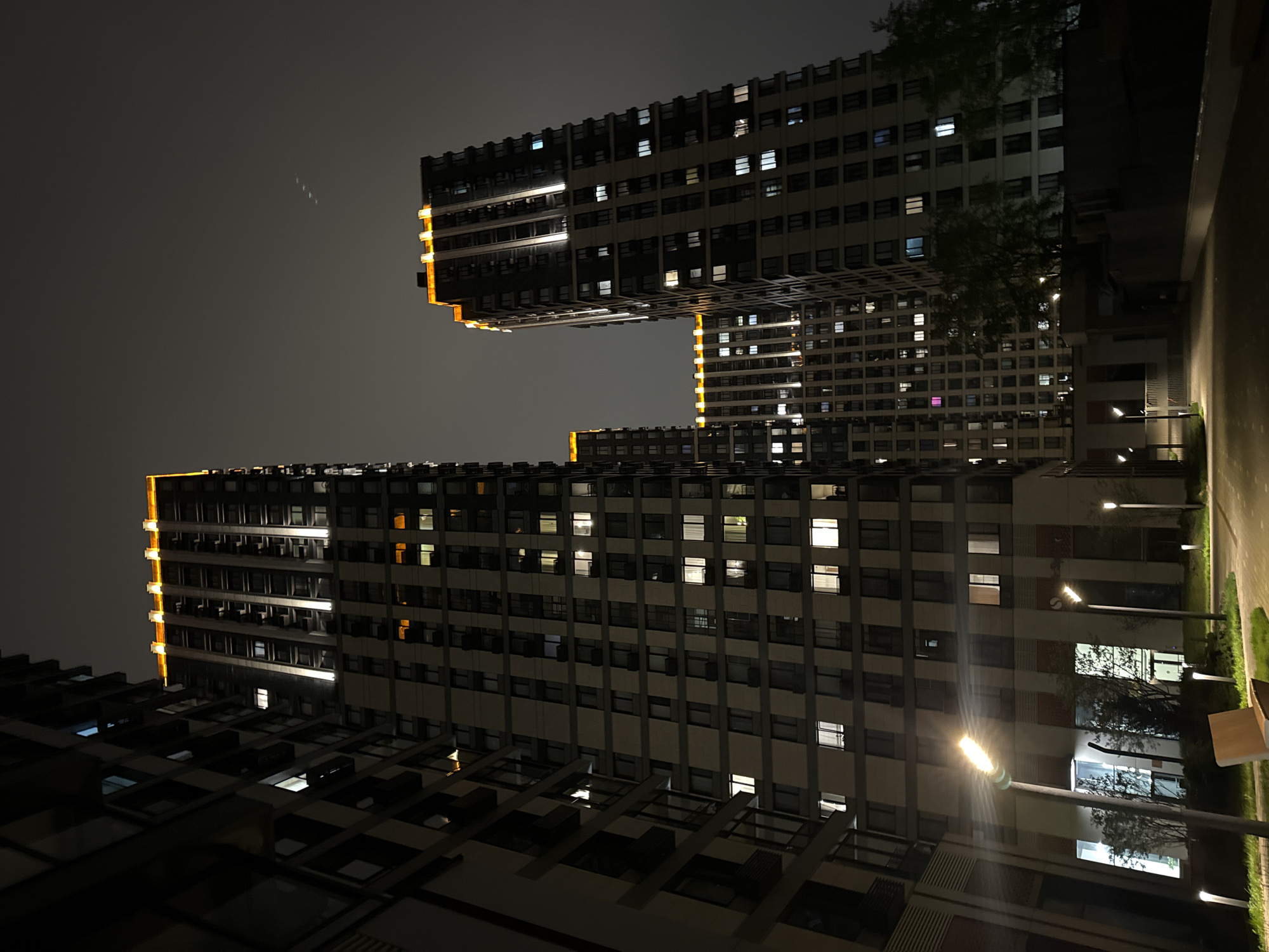 The image shows a low-angle view of illuminated apartment buildings at night. The buildings are located in Moscow, Russia, specifically in the Butyrsky district. The architecture features large residential towers with lit windows and distinctive vertical lighting accents at the edges. The sky is dark, and the ground area near the buildings is dimly lit by street lamps. Some trees are visible near the base of the buildings. There are no people visible.