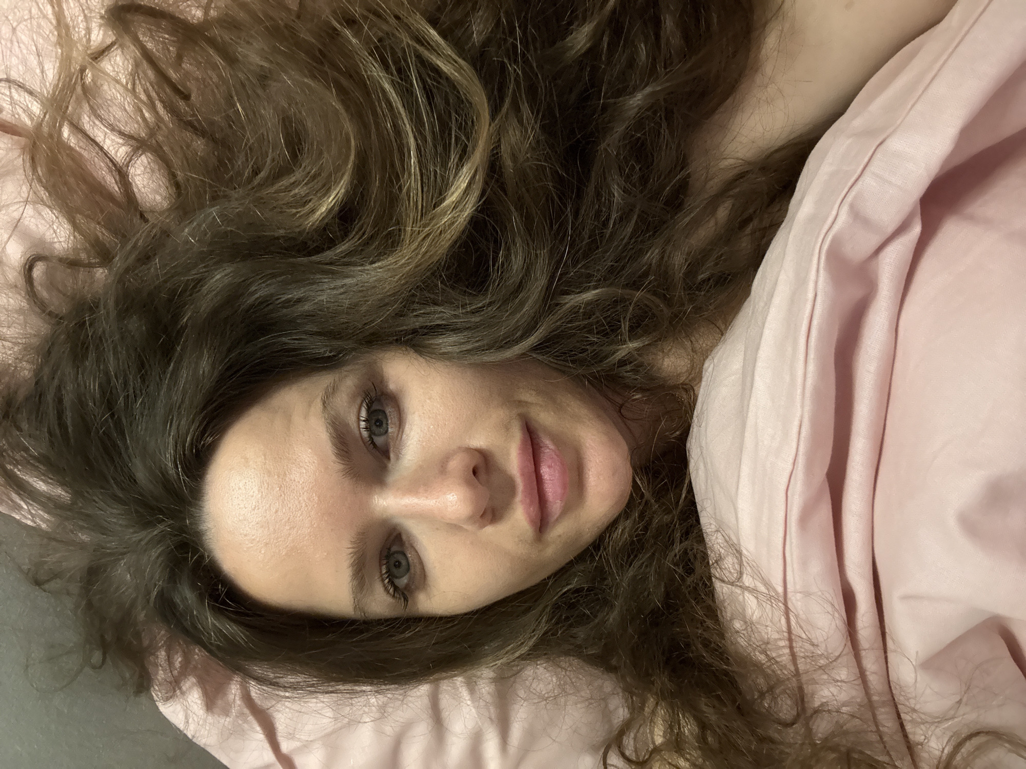 A woman lies in bed, looking directly into the camera from a slightly overhead perspective. Her long, wavy, brown hair, with subtle lighter highlights, is spread out around her head on a soft pink pillow. Her blue-gray eyes are open and focused, and her lips curve into a gentle, faint smile. A pink duvet or blanket covers the right side of her body, obscuring her shoulders and torso, suggesting she is under the covers. The lighting is soft and even, typical of an indoor setting, likely a bedroom. The image conveys a sense of calm and intimacy. There are no visible location cues within the image to indicate Moscow, Russia, nor any discernible text. The time of day cannot be determined from the indoor lighting.