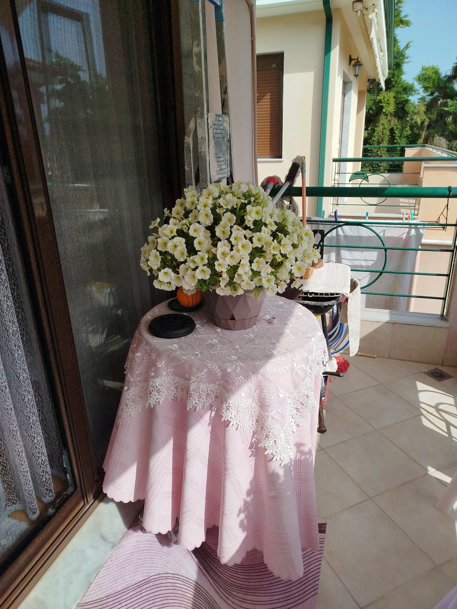 A round table covered with a pink tablecloth decorated with white lace is set on a balcony in Sithonia Municipal Unit, Greece. A large pot of white petunias sits on the table, accompanied by an orange decorative object that resembles a mini basketball. To the left of the table is a large glass door with a screen, and beyond that, a curtain. The balcony floor is tiled, and a rug with purple stripes lies in front of the table. In the background, a railing separates the balcony from the exterior. A chair with a folded towel on it is visible behind the table. The scene is illuminated by sunlight casting shadows on the floor.